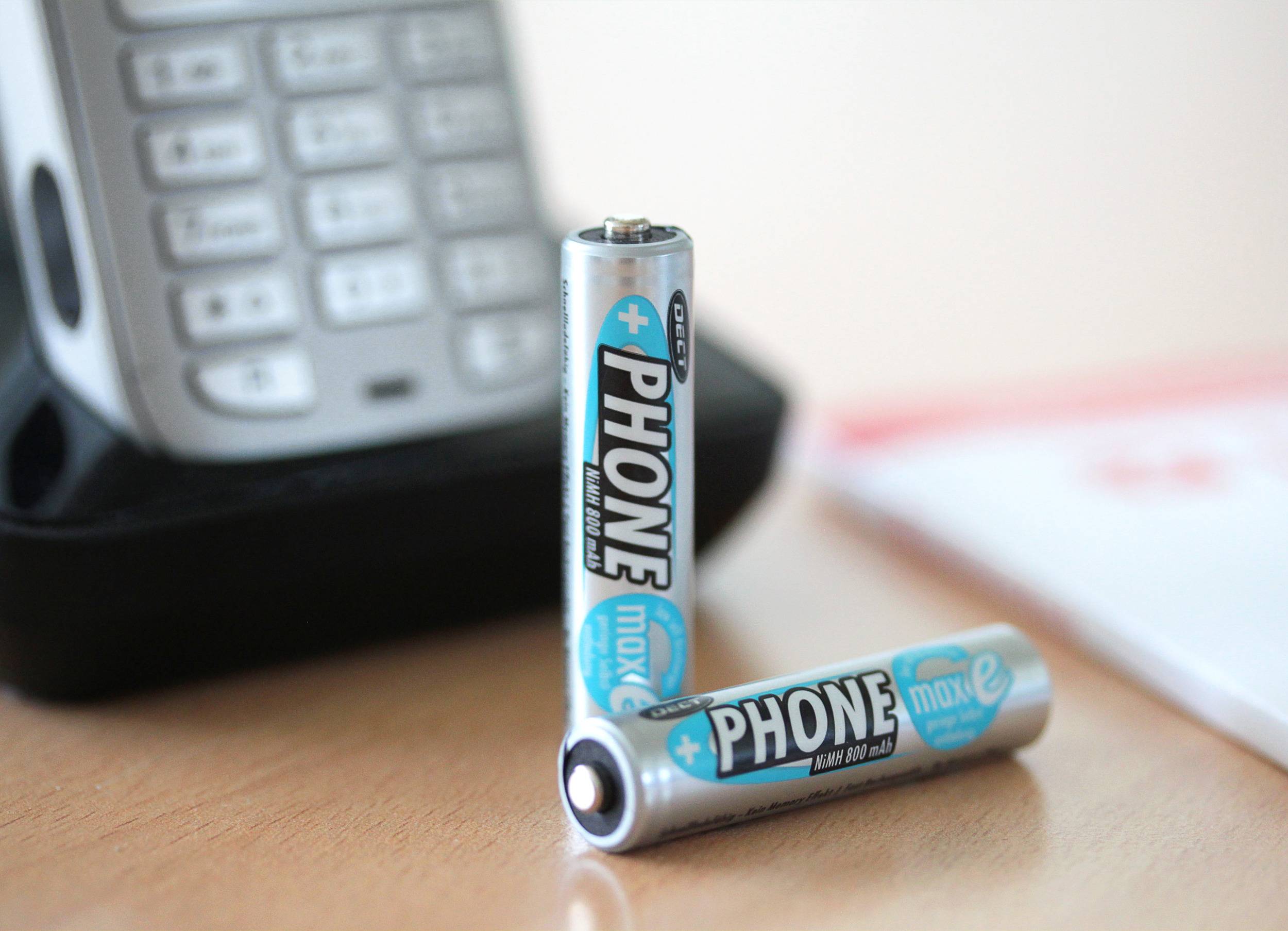 Two AA batteries labelled 'PHONE max e' are lying in front of a cordless telephone on a table.
