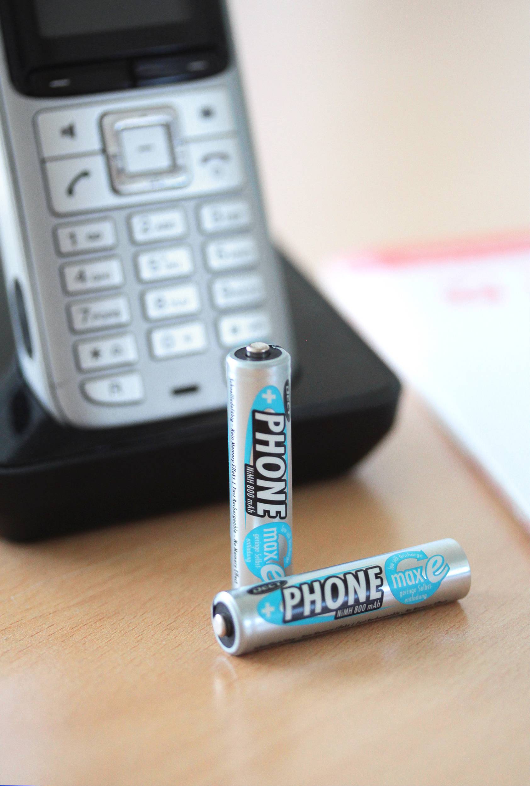 Two batteries with the label 'PHONE max e' are lying in front of a cordless telephone.
