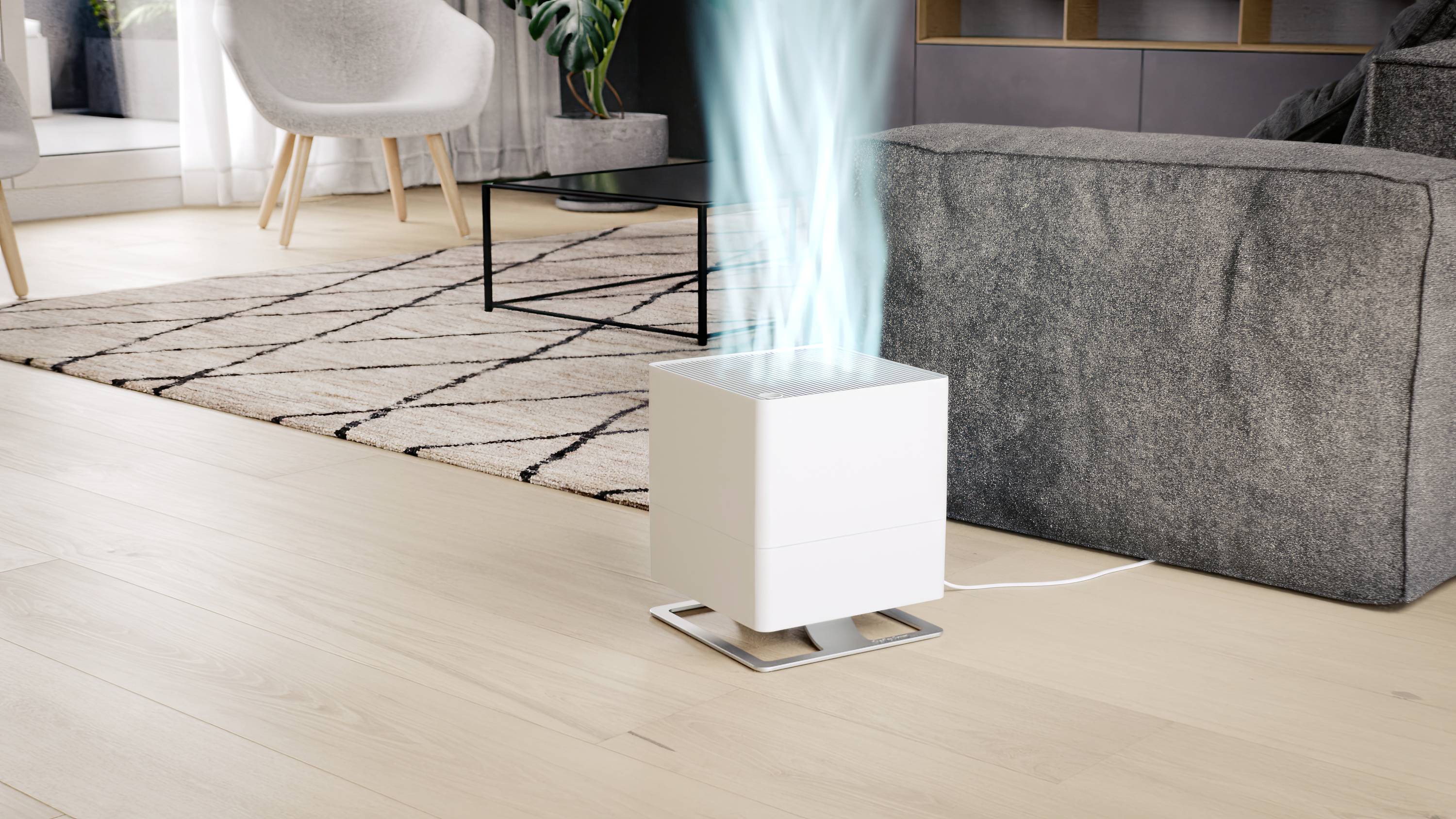 A white air purifier stands on a wooden floor next to a grey sofa. Blue vapour rises from the device.
