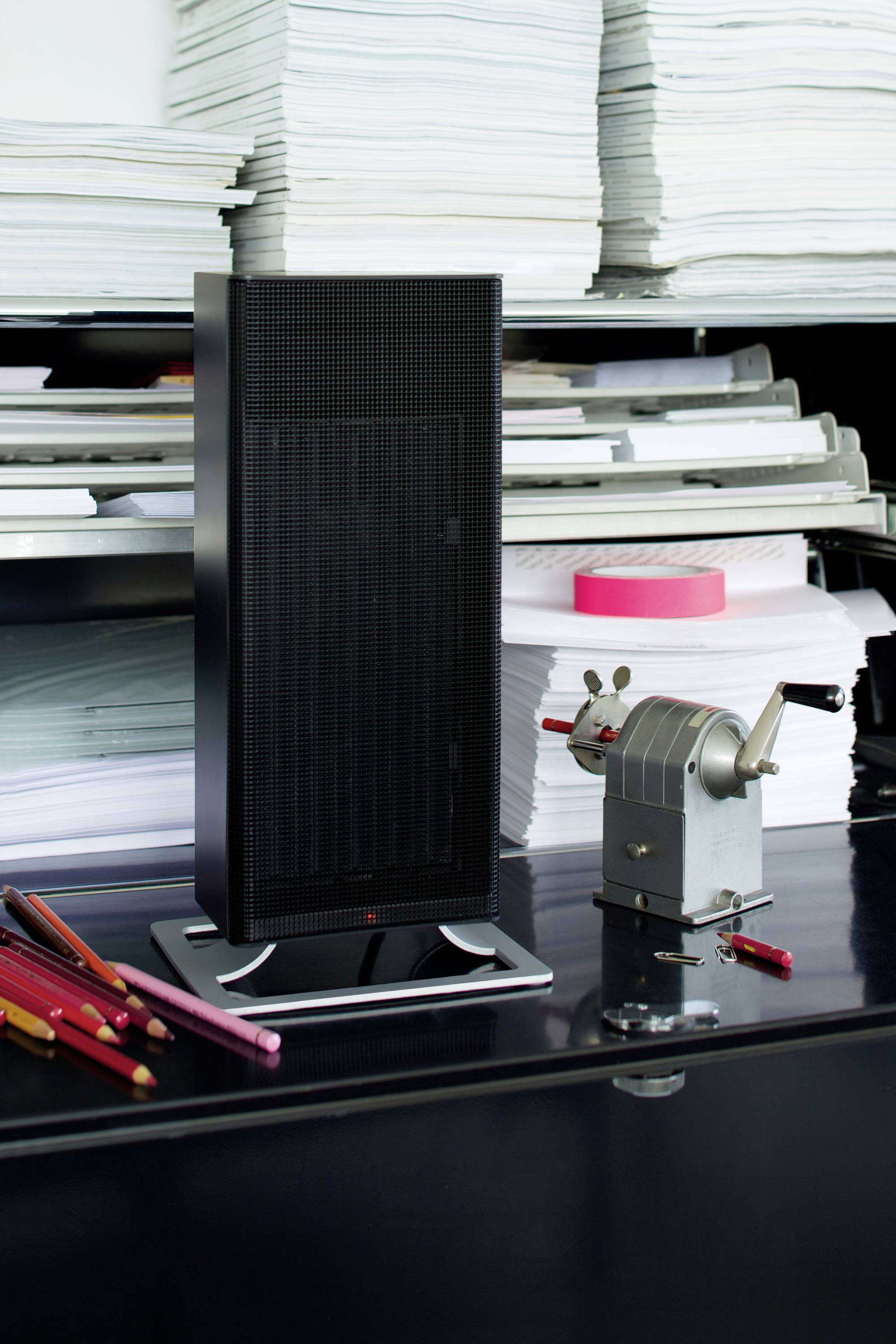A black tower fan stands on a table covered with stacked papers. Next to it lie pencils and an old metal pencil sharpener.