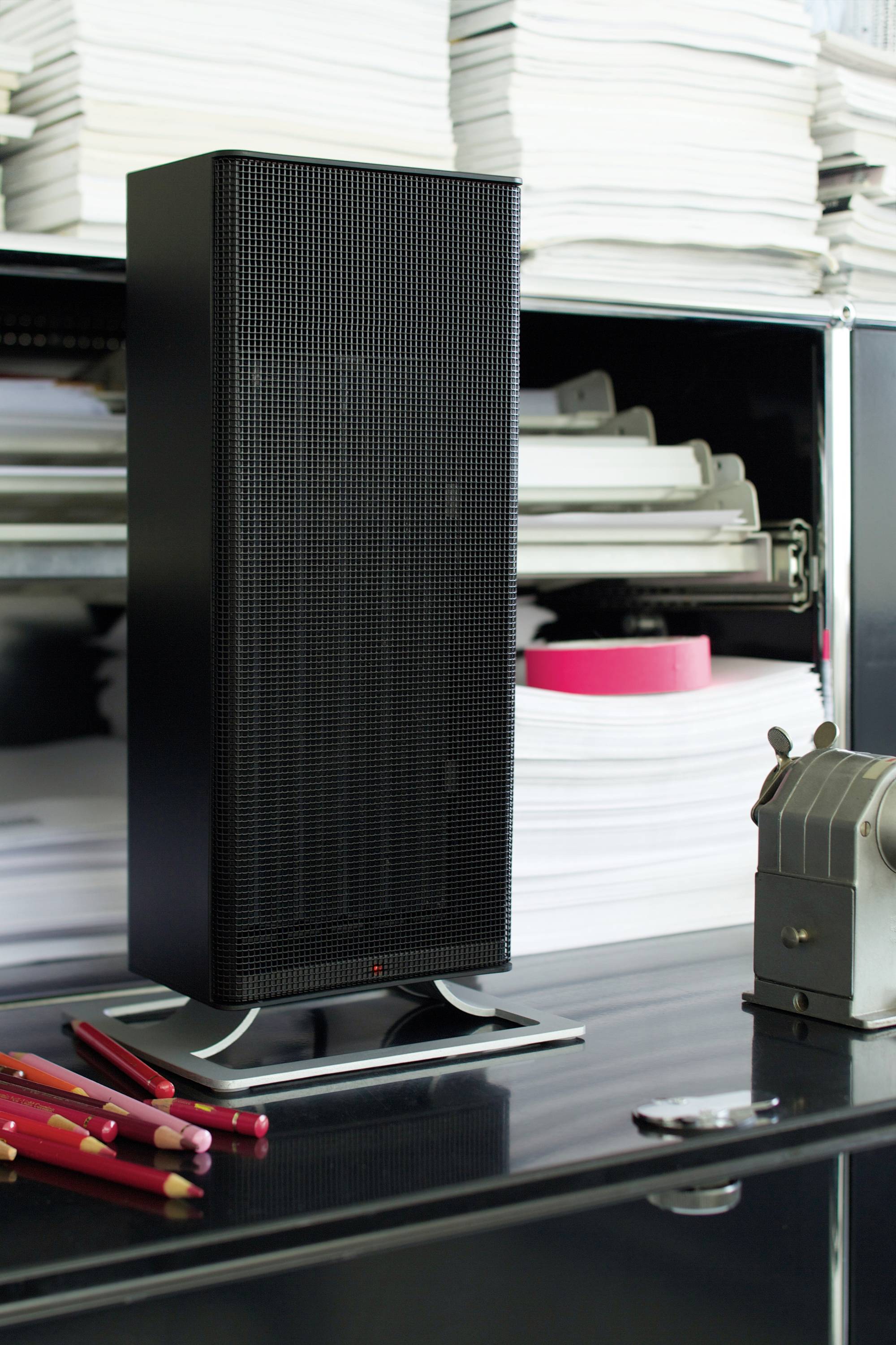 Black speaker stands on a table in front of a shelf with stacked paper piles; red pens and a pencil sharpener lie beside it.