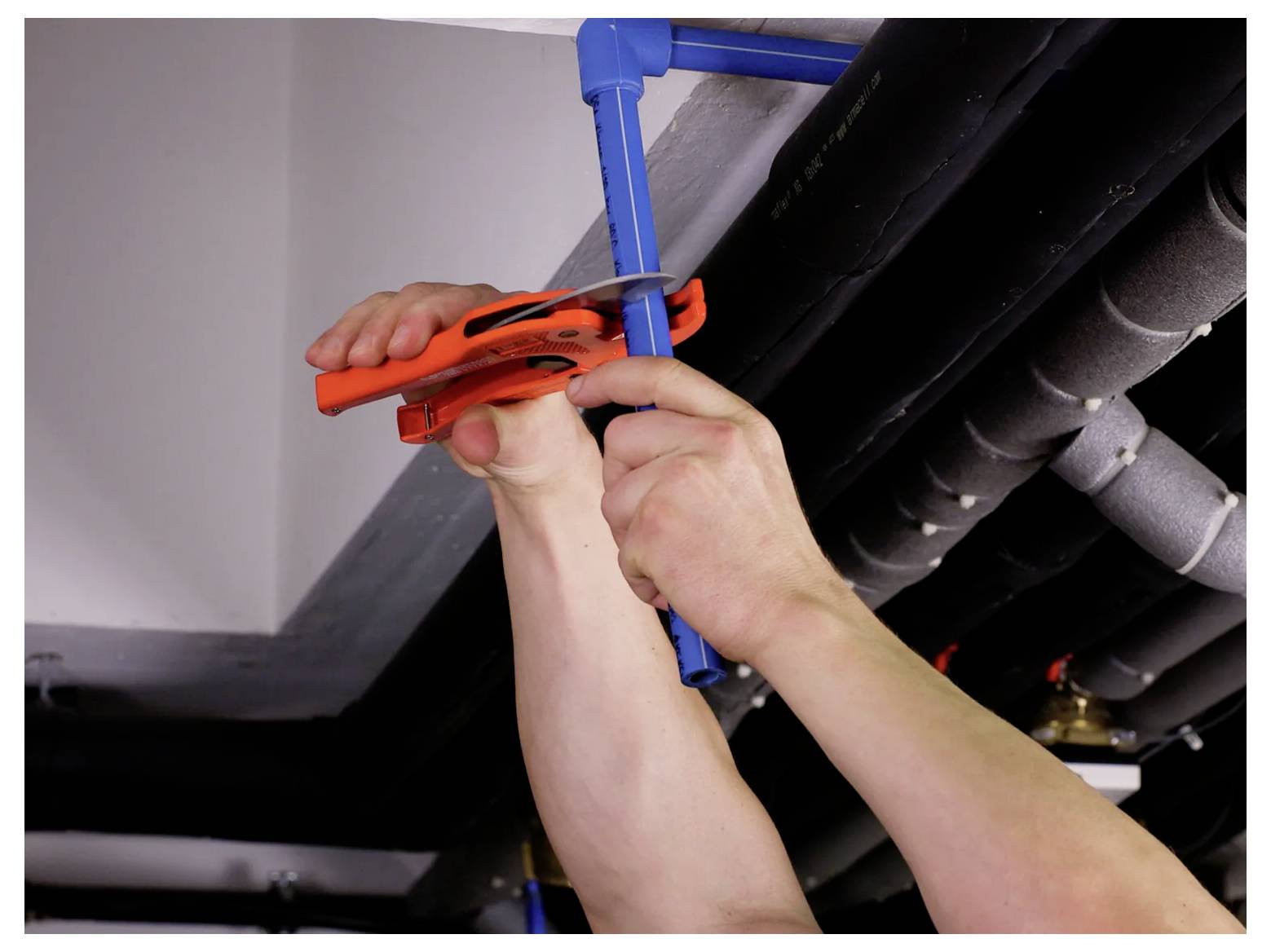 Close-up of hands using a pipe cutter to cut a blue pipe under a ceiling, suggesting plumbing or installation work.