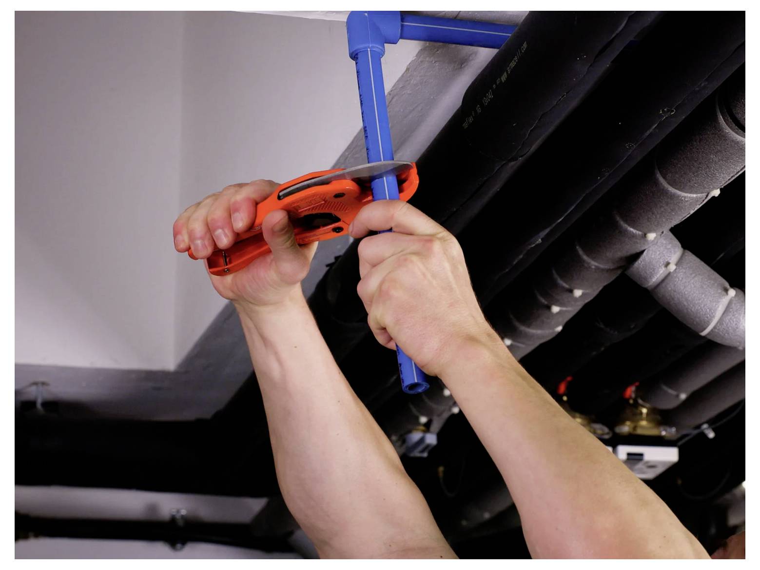 Hands using an orange pipe cutter to cut a blue plastic pipe beneath ceiling plumbing.