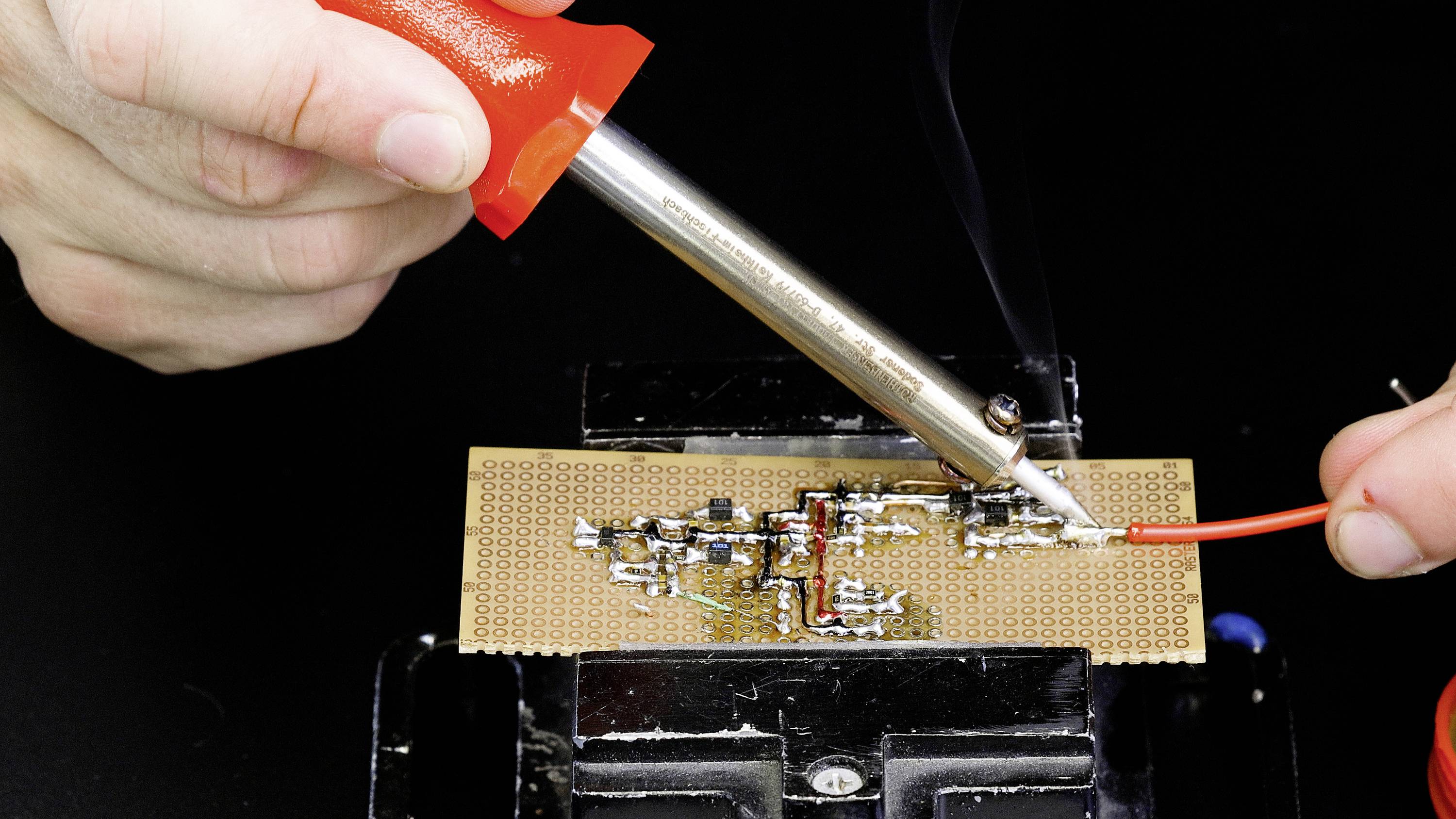 A person is soldering with a soldering iron and a red cable on a circuit board. Manufacturing or repair of electronic circuits.