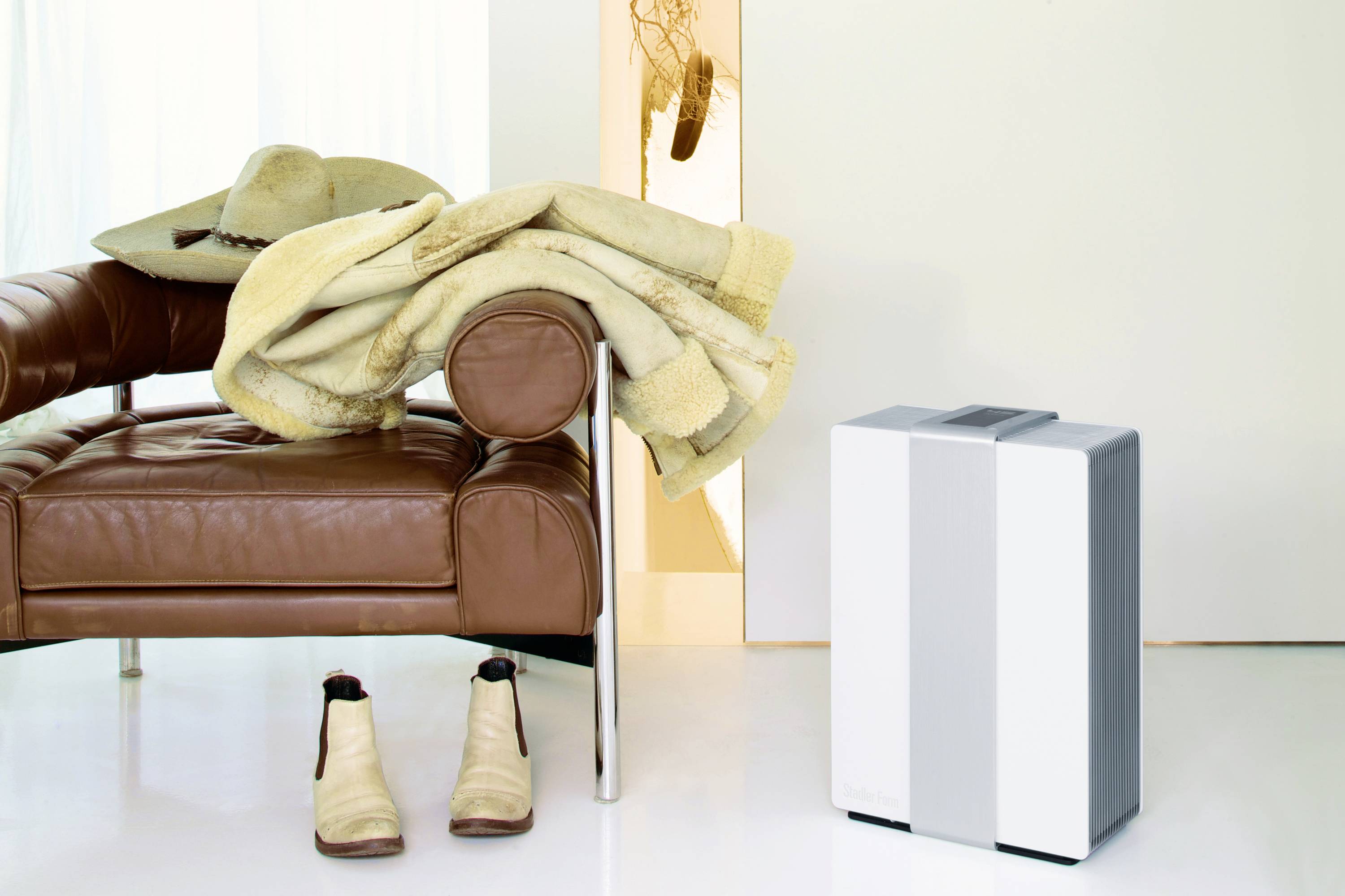A brown leather armchair with a hat and coat draped over it. Next to it stand cowboy boots. To the right, a modern air purifier sits on a white floor.