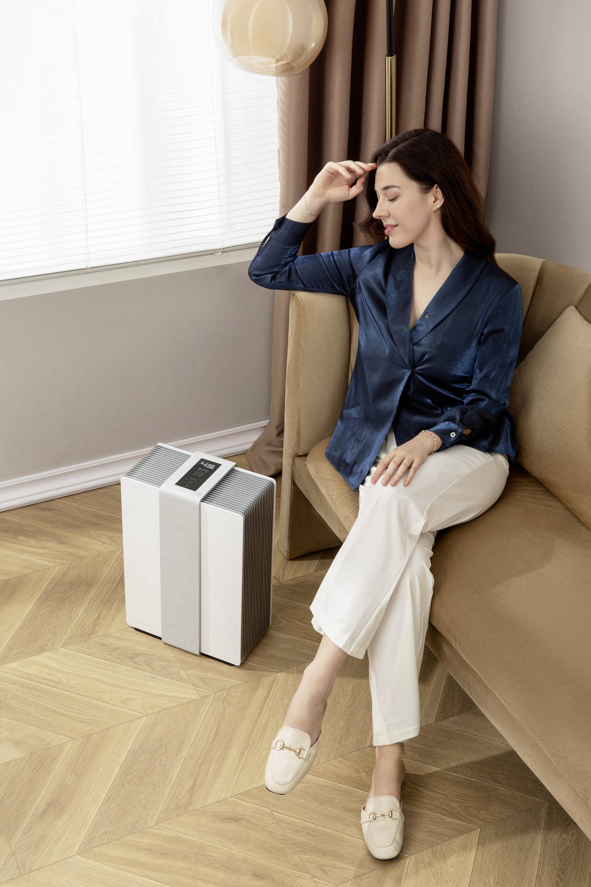 A woman wearing white trousers and a blue blouse is sitting on a sofa next to an air purifier. She gently touches her hair and looks relaxed.
