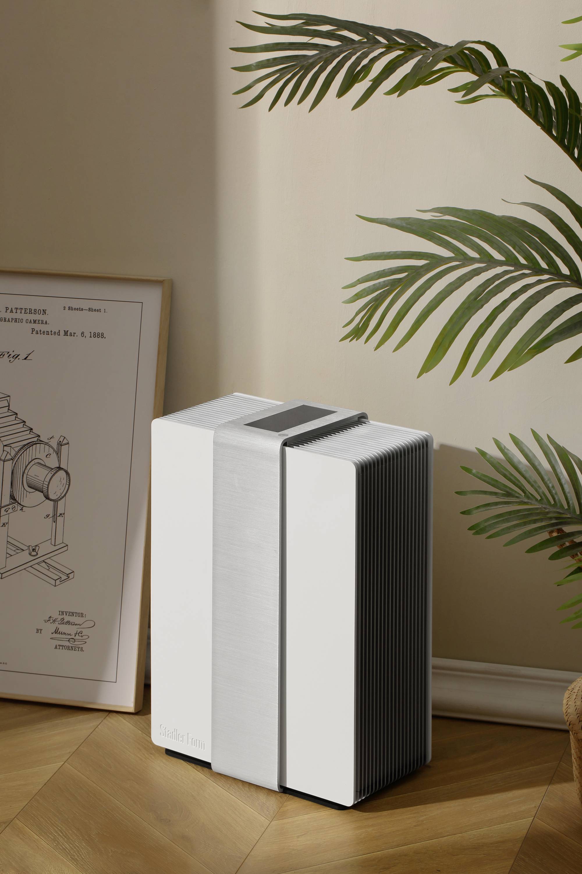 A modern air purifier stands on a wooden floor, next to a plant and a framed technical poster. The design is minimalist.