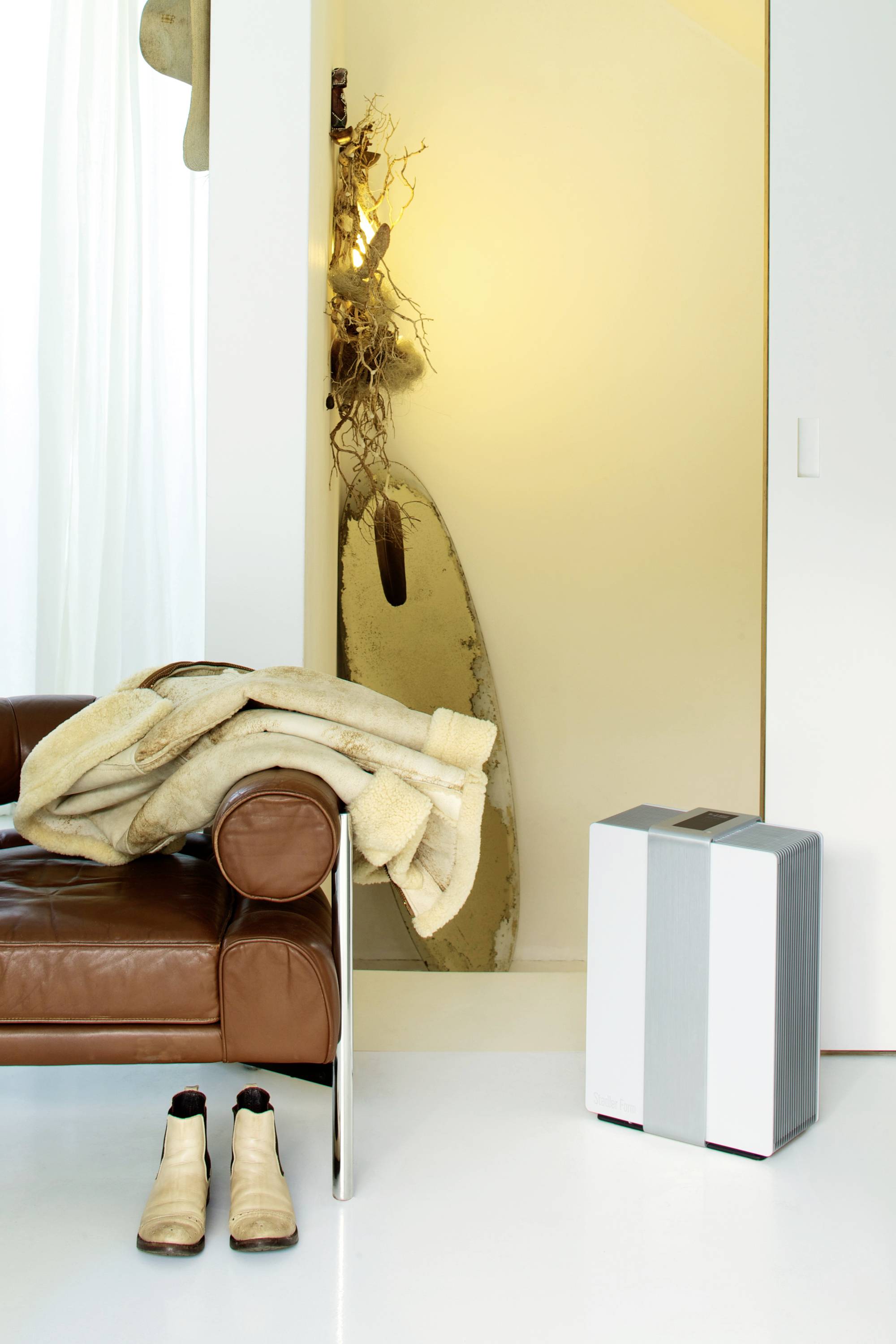 A modern furnished living room with a leather armchair, fur jacket, discarded boots and an air purifier on a white floor.