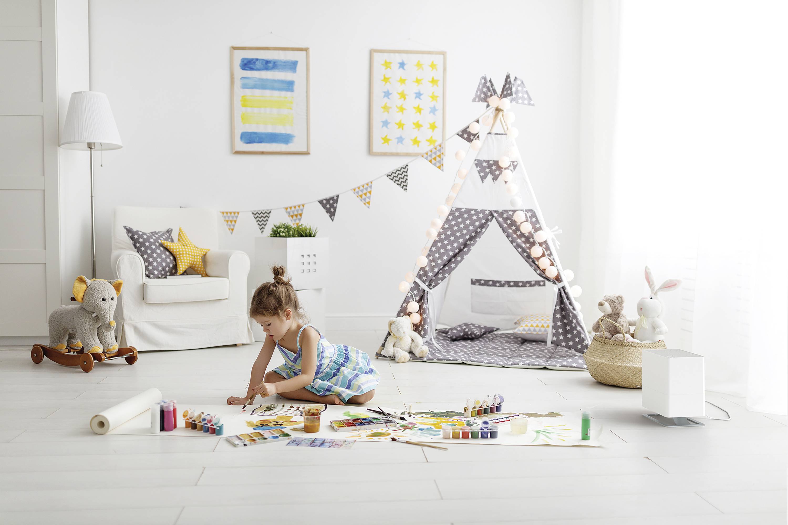 A little girl is drawing on the floor of a bright children's bedroom, surrounded by toys and a decorative teepee tent.
