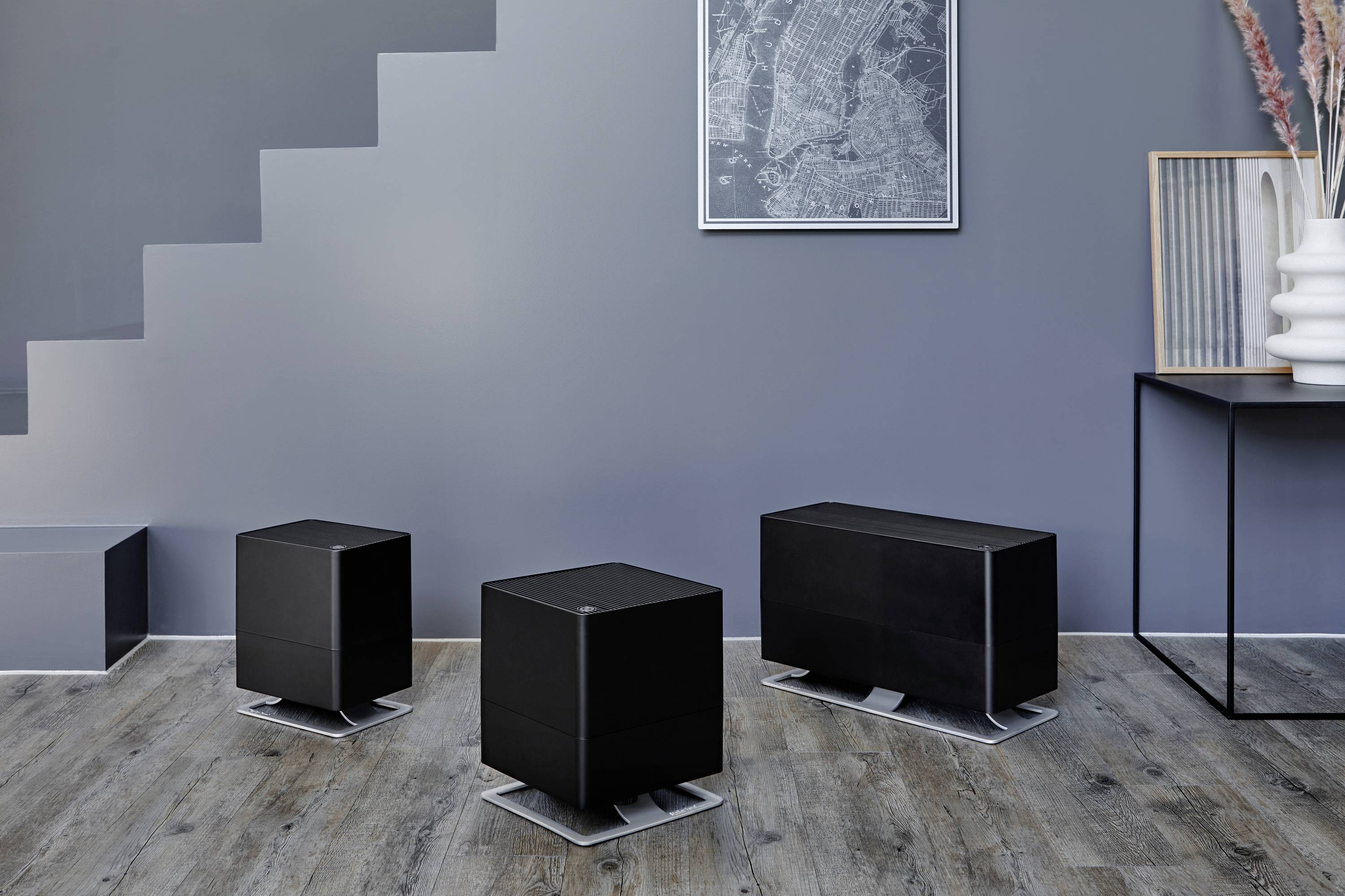 Three black air humidifiers in various sizes on a wooden floor in a modern room with a grey wall and minimal decoration.