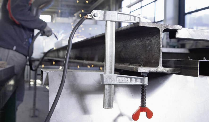 Worker in factory hall, welding a metal beam. Sparks flying. Metal clamp holds workpiece firmly.