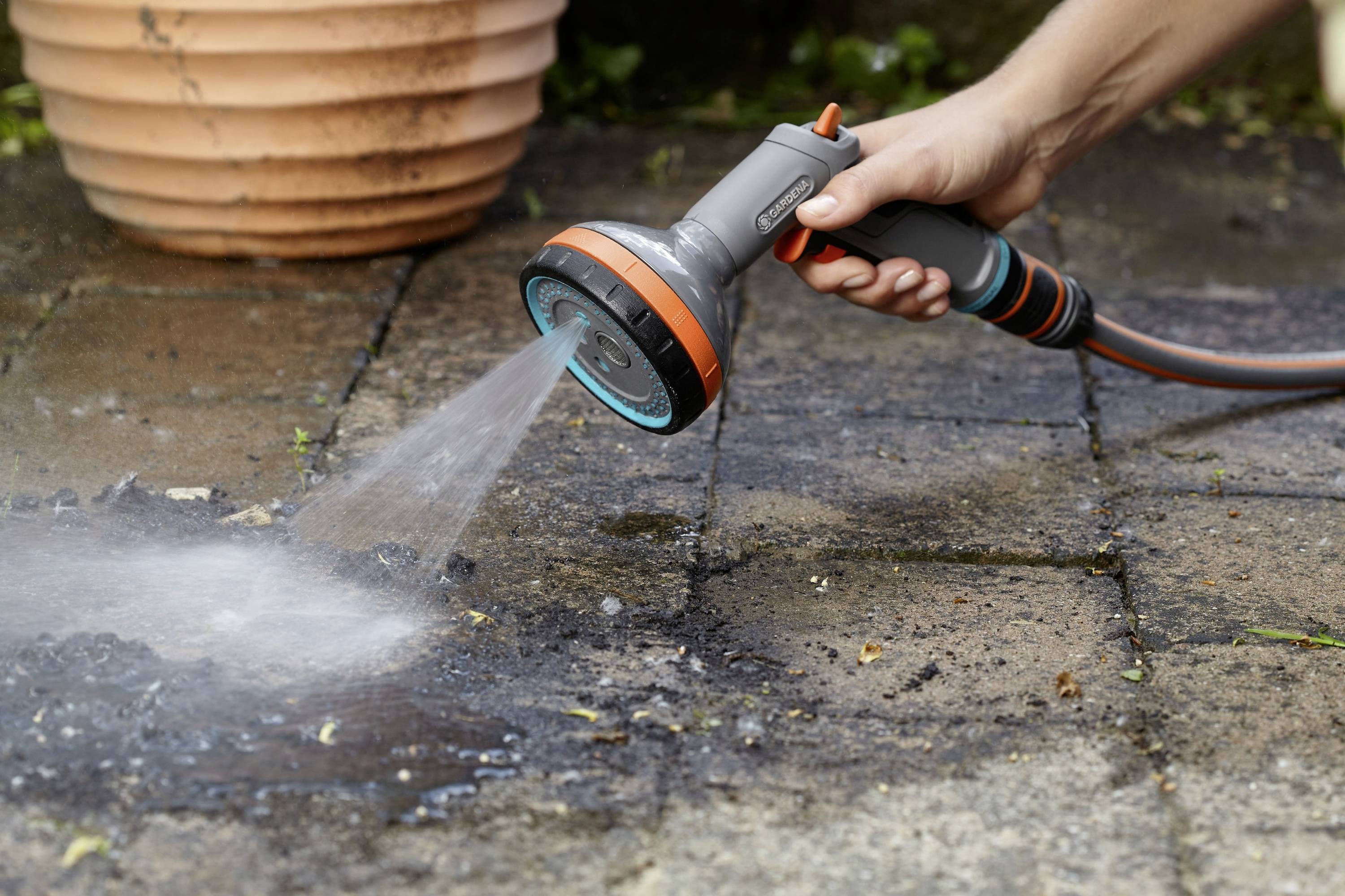 A hand is holding a garden hose spray nozzle and spraying water onto a wet, paved terrace surface next to a flowerpot.