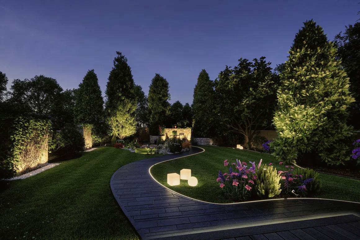 A beautifully lit garden at night with a lawn, trees, flowers and a winding path threading through the scene.
