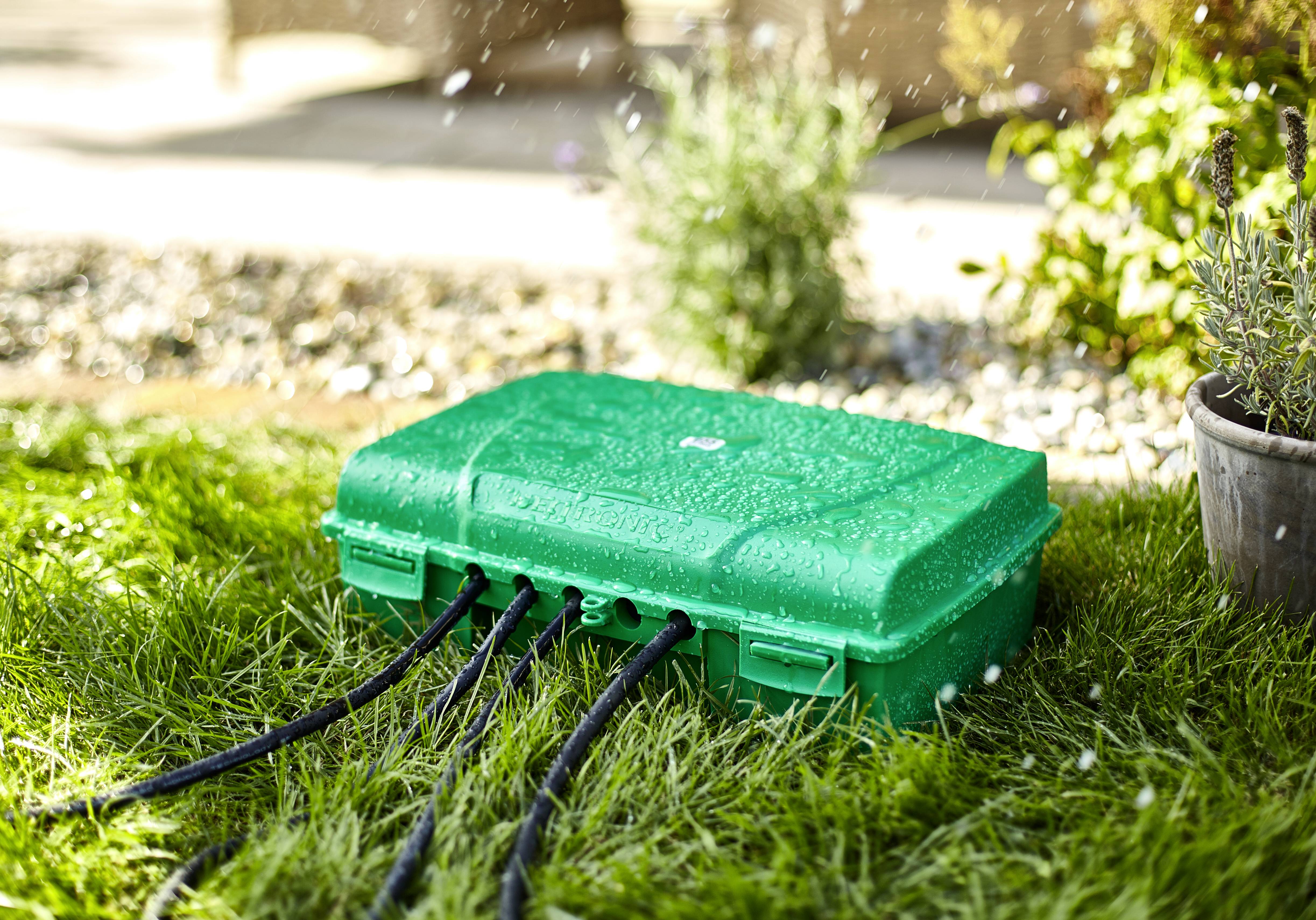 A green cable box sits on grass, protecting cables from moisture. It is being sprayed with water and stands next to a flowerpot.