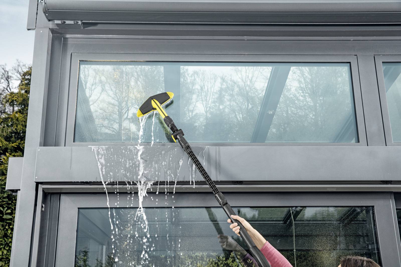 A person uses a long-handled tool with a cleaning attachment to wash a large glass window on a building, with water visibly splashing.