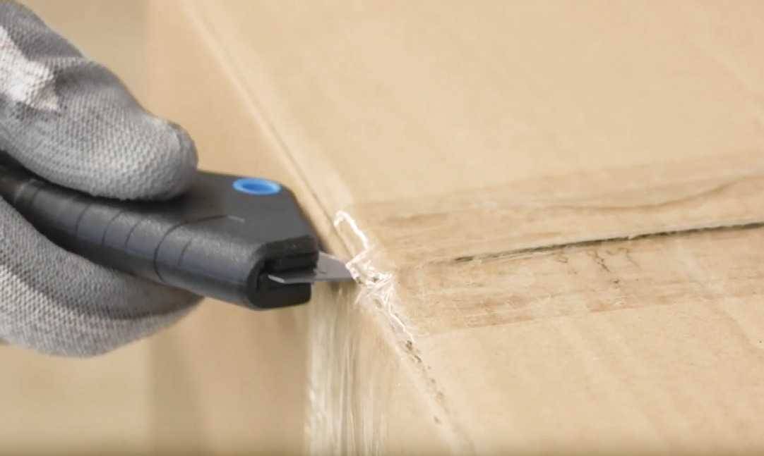 A hand wearing a glove holds a black safety knife and cuts open a cardboard box.