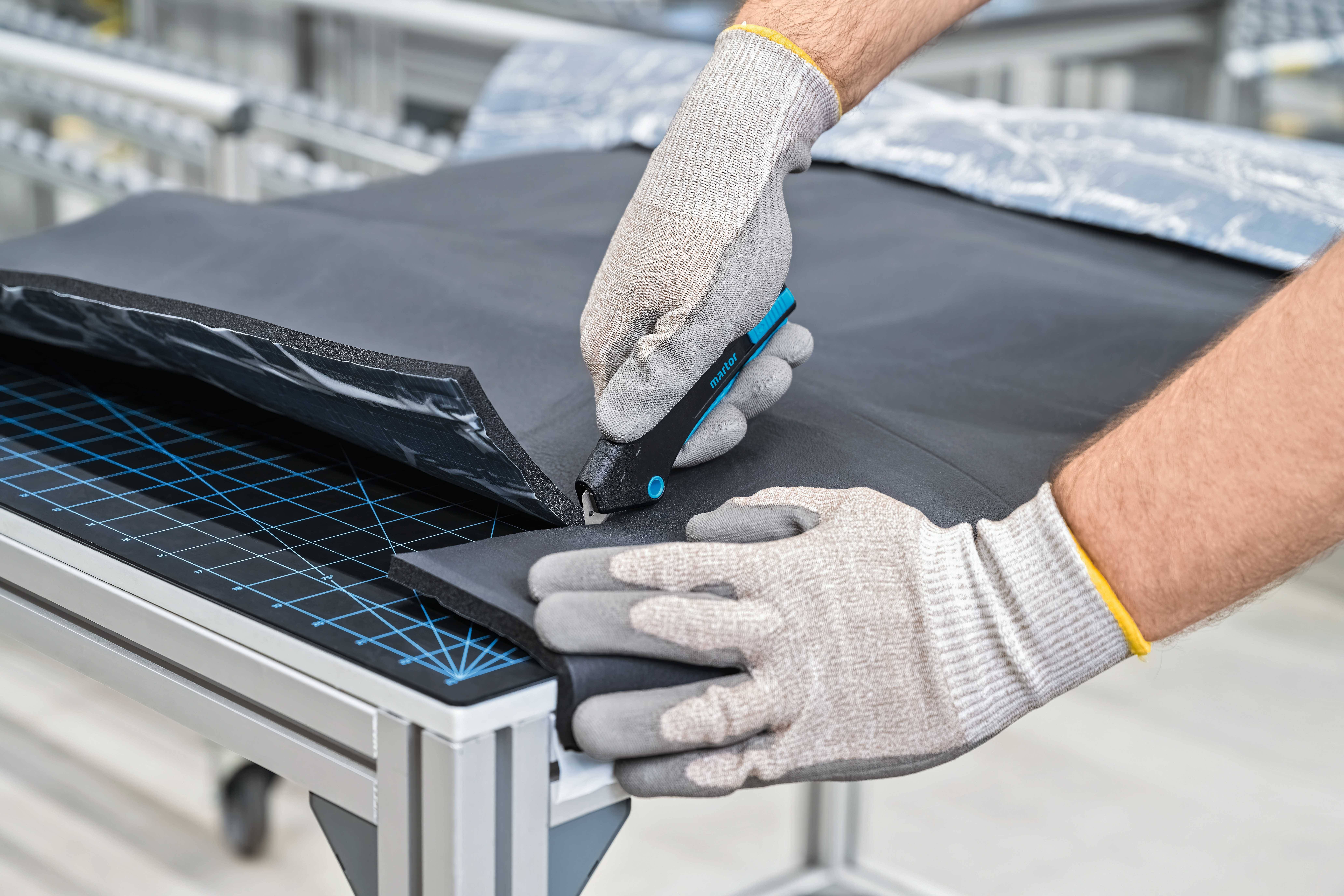 A person is cutting a black material with a craft knife on a cutting table, wearing work gloves.