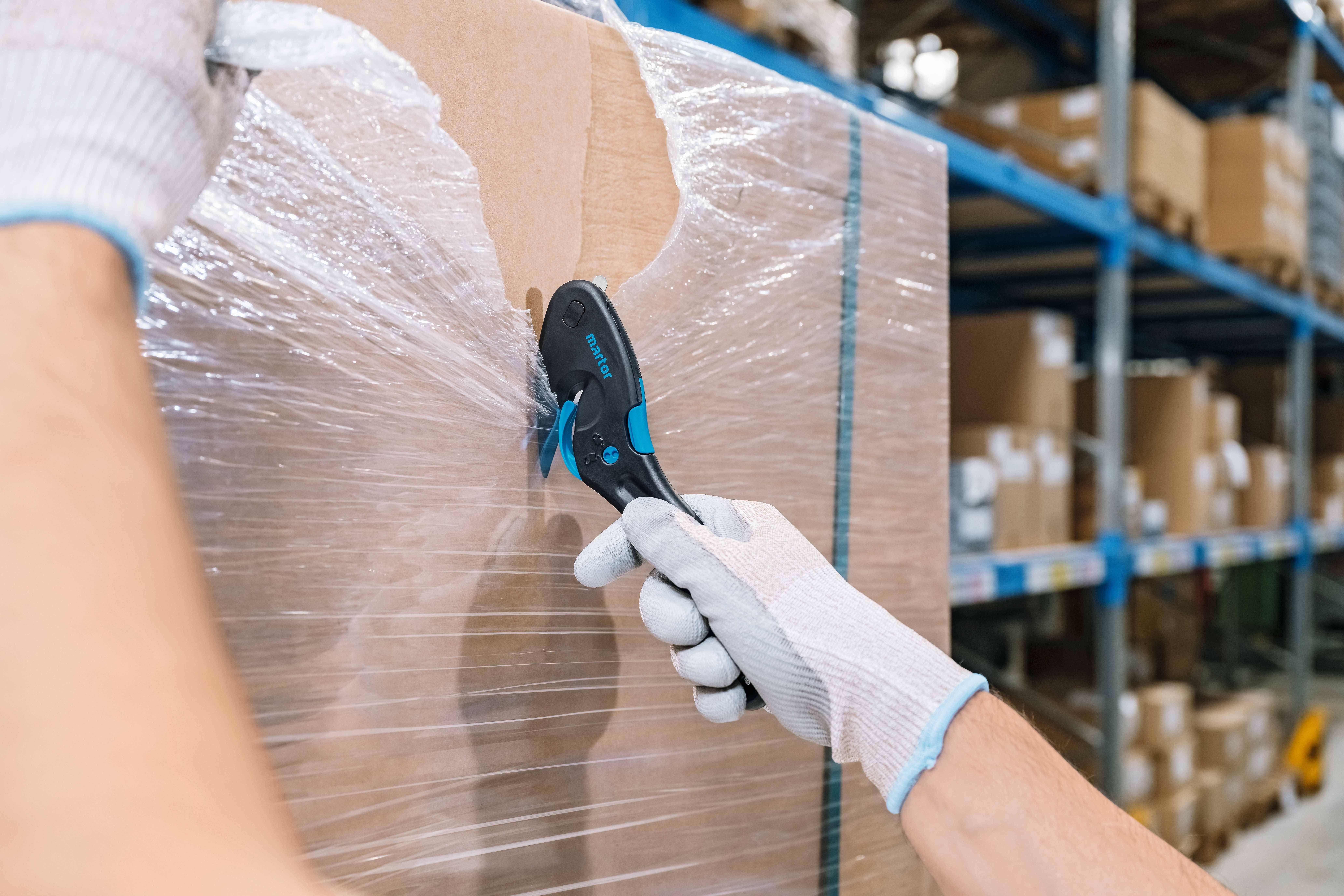 A person wearing gloves is opening a cardboard box wrapped in film using a black safety knife. Warehouse shelving can be seen in the background.