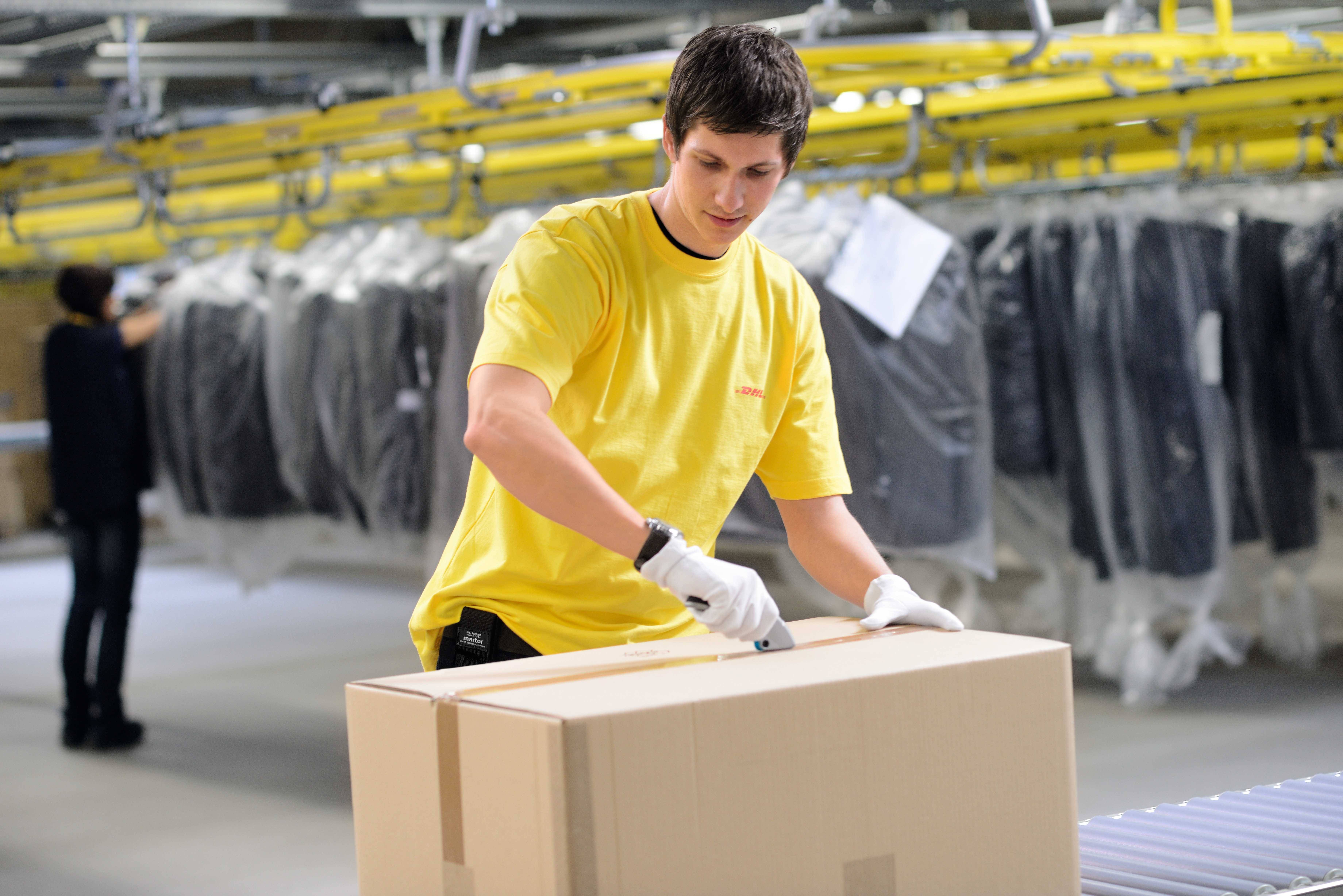 A staff member in a yellow T-shirt opens a large cardboard box with a utility knife in a warehouse, with clothing racks visible in the background.