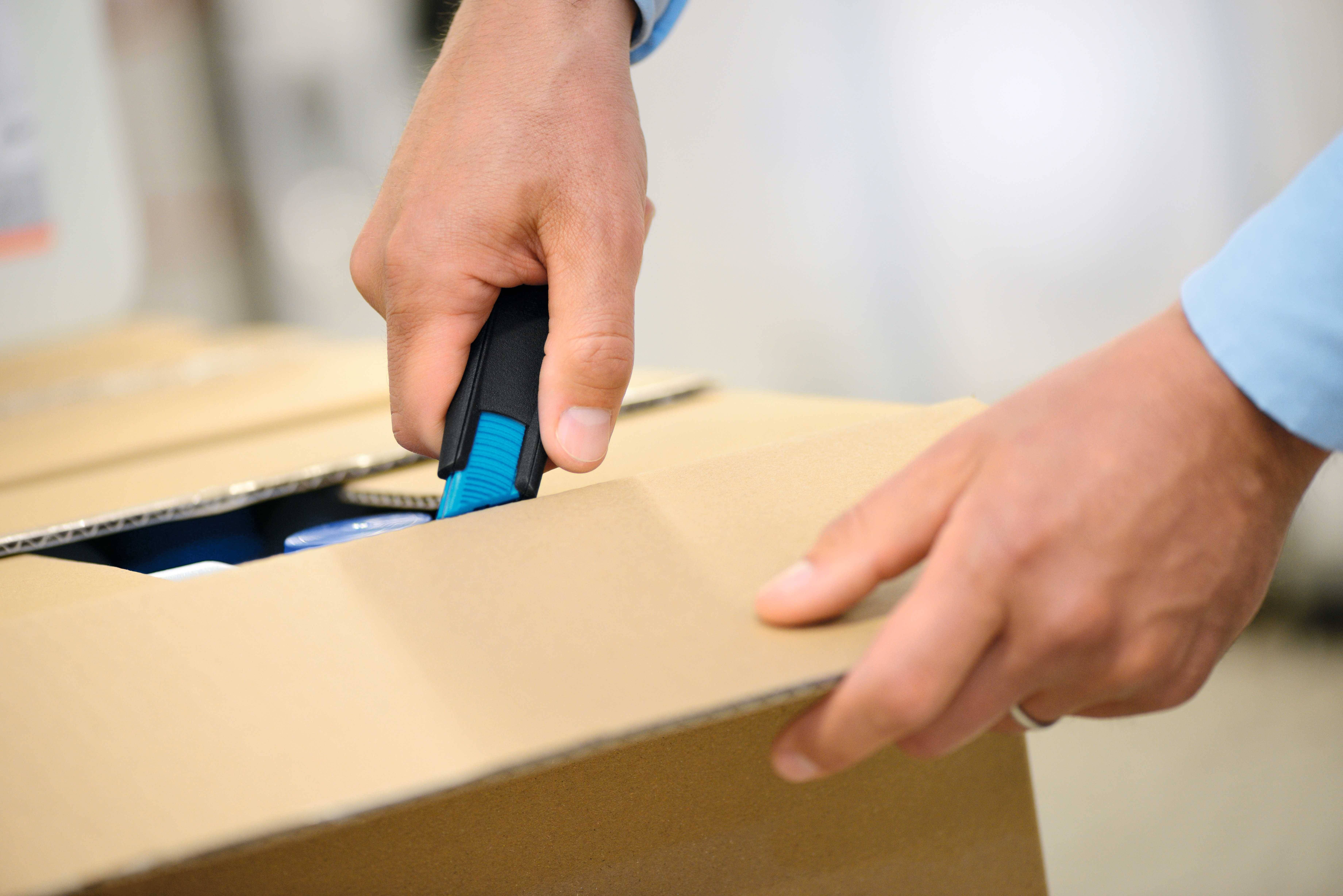 A person opens a cardboard box using a Stanley knife. Hands in focus, details of the box and knife visible.
