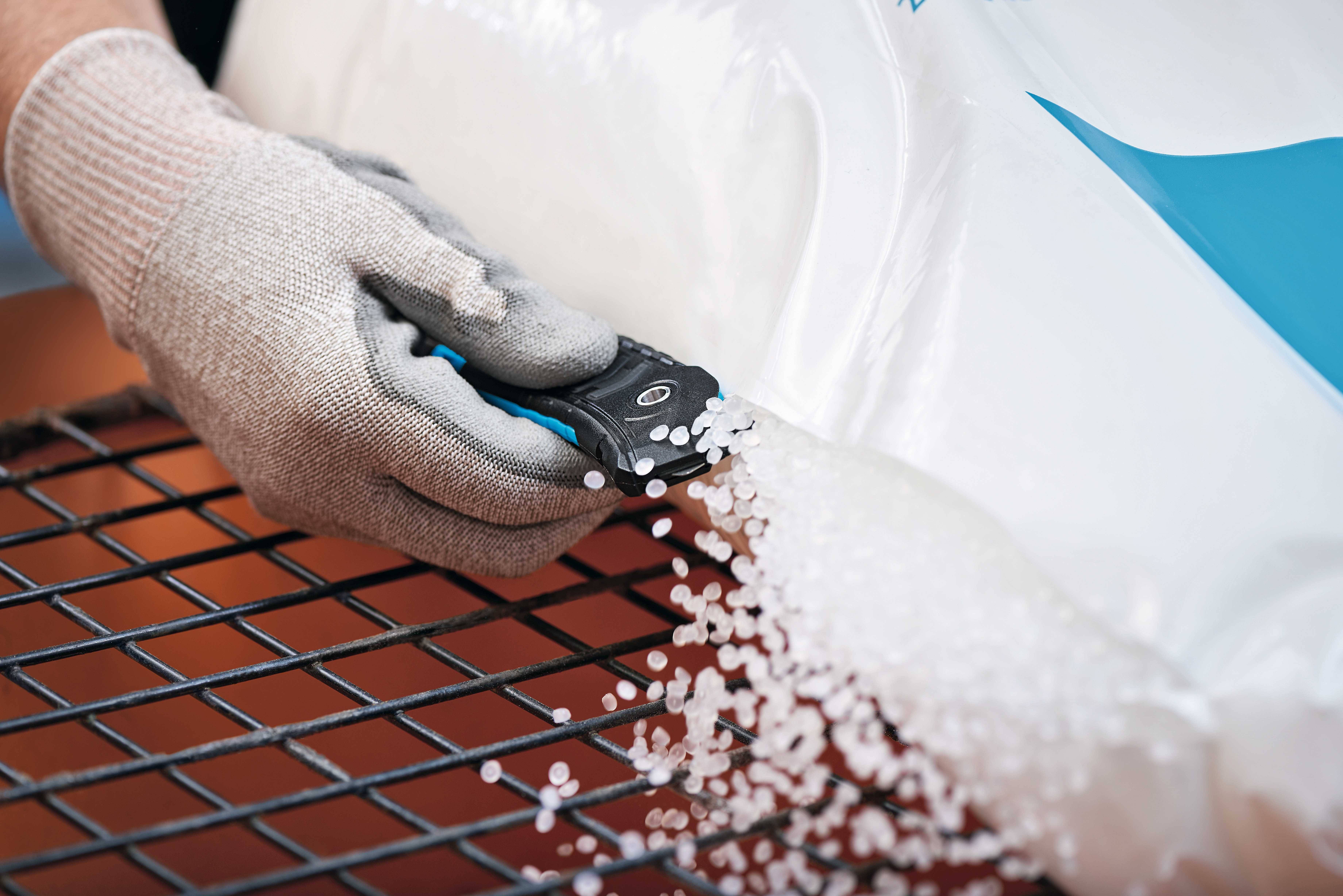 A gloved hand cuts open a plastic bag, from which white plastic pellets cascade onto a grid.