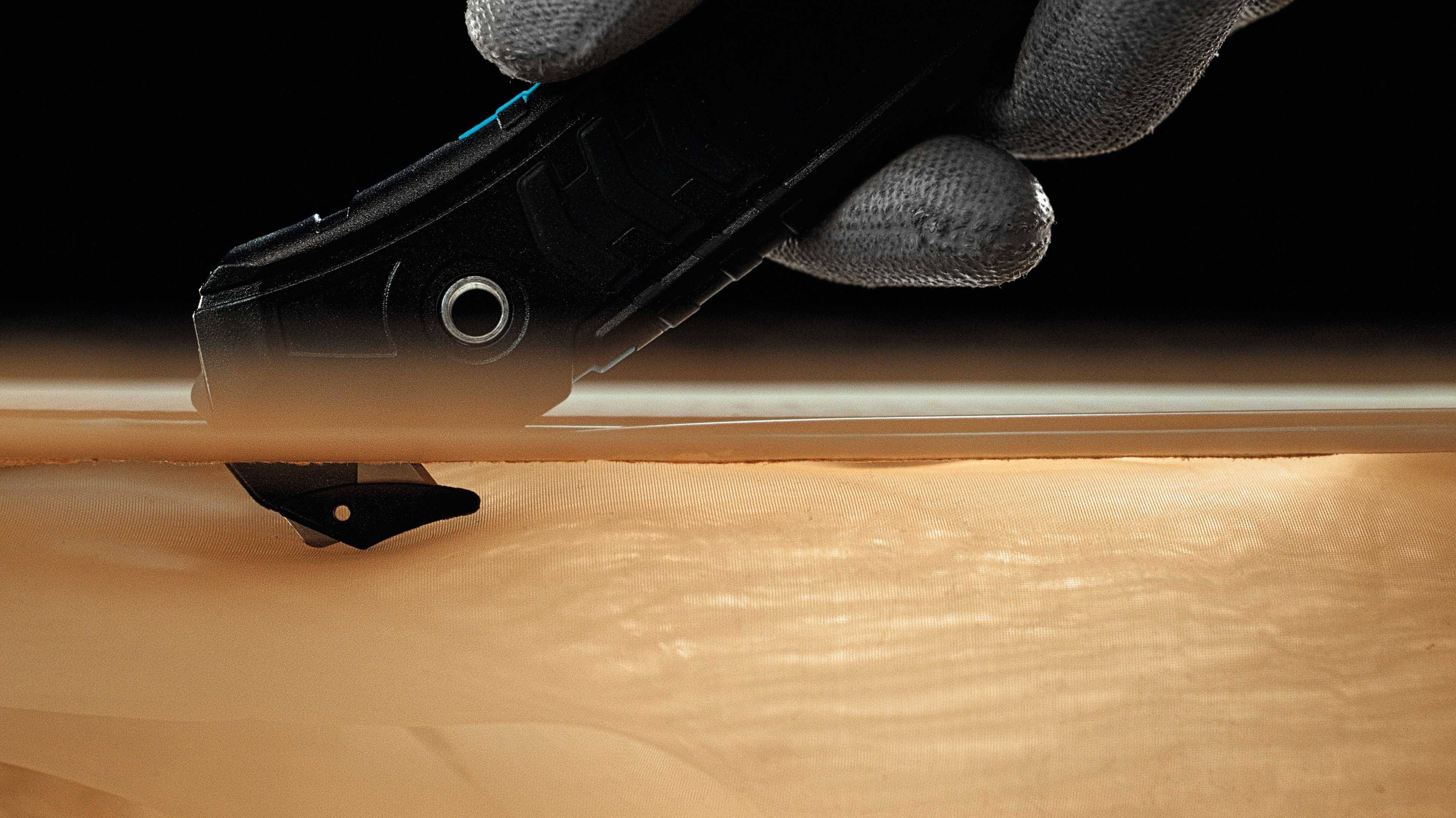 A hand wearing a grey glove cuts a smooth, matte surface with a black utility knife.