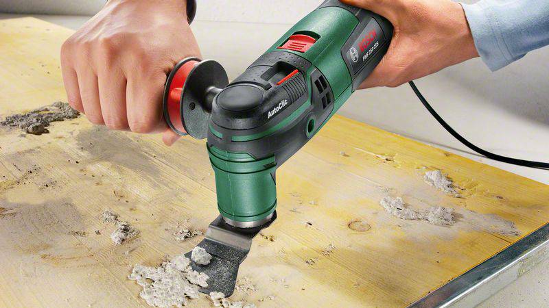 A hand is holding a green multi-tool removing mortar from a wooden board.
