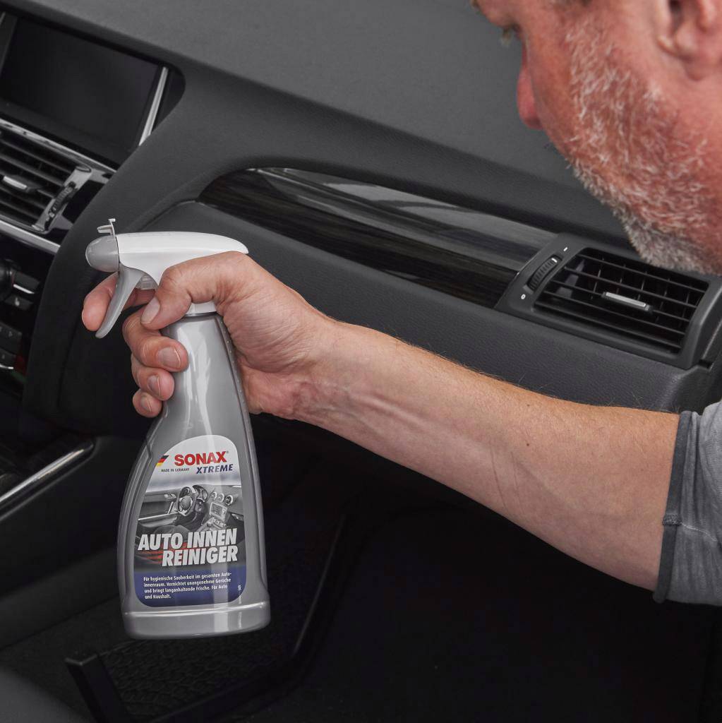 A person is spraying 'Car Interior Cleaner' onto the dashboard of a car. Product by Sonax for cleaning the vehicle's interior.