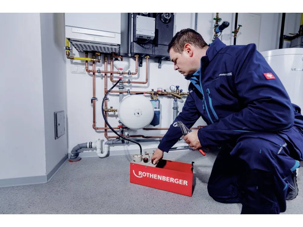 A technician wearing a blue uniform kneels by a heating system, using a tool from a 'Rothenberger' box, inspecting pipes and gauges.