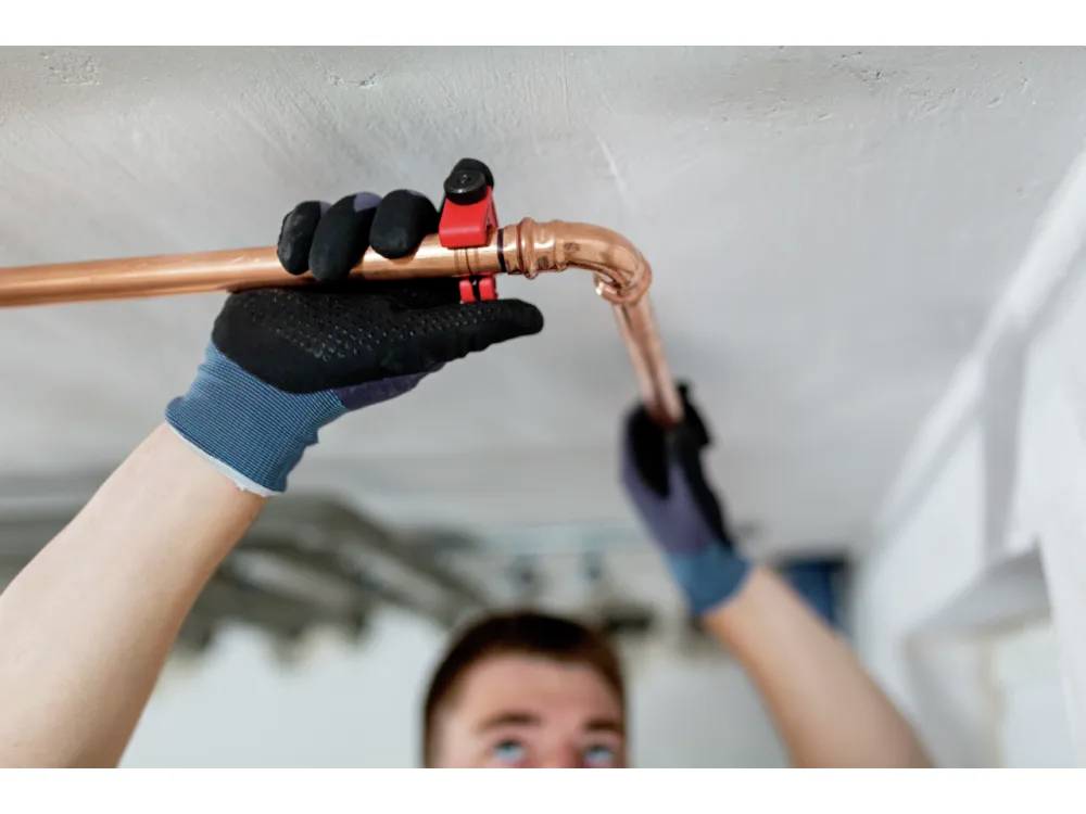 A person using a pipe cutter to fix copper plumbing under a ceiling, wearing protective gloves.