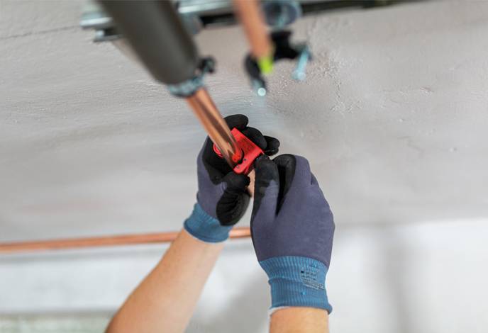 A person wearing gloves is fixing a copper pipe on a ceiling, using a wrench or similar tool, focusing on plumbing repairs.