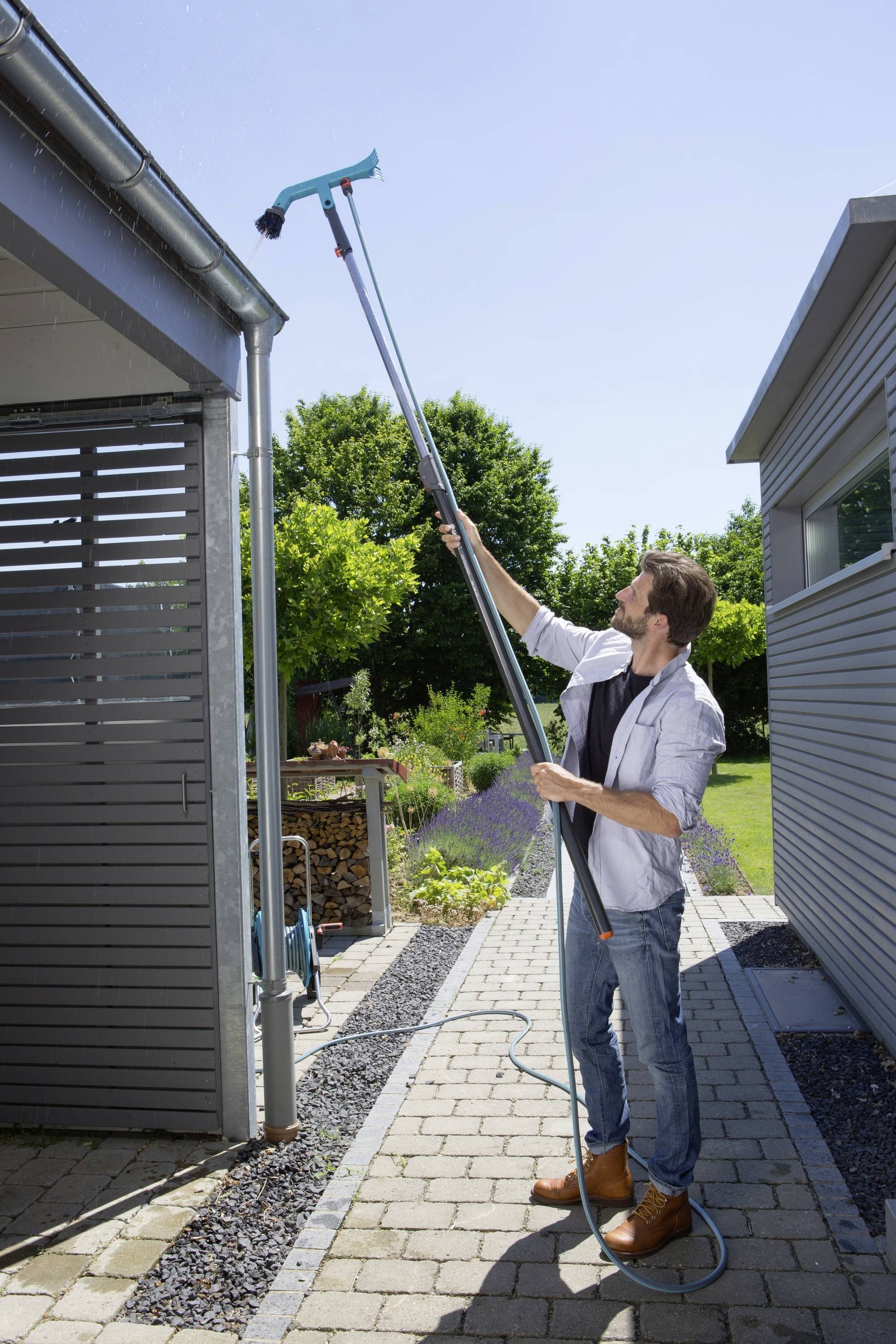 A man is cleaning a guttering of a house with a long telescopic pole in a sunny garden.