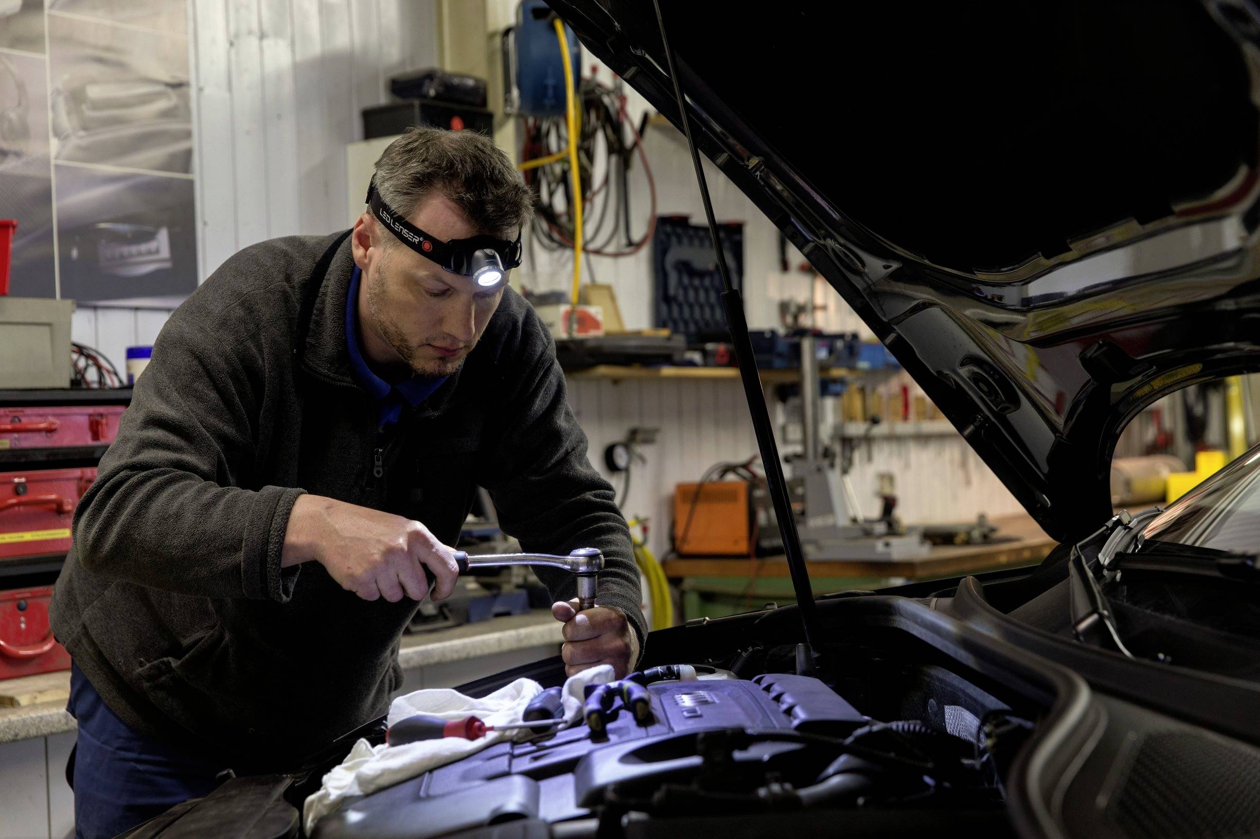 Ein Mechaniker arbeitet mit einer Taschenlampe am offenen Motor eines Autos in einer Werkstatt. Werkzeuge und Ausrüstung sind im Hintergrund sichtbar.