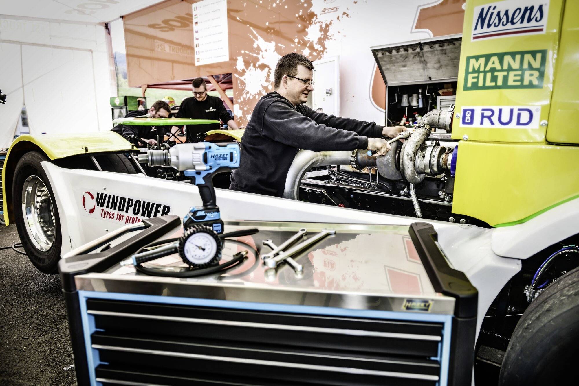 A man is working on a racing truck engine in a workshop, surrounded by tools and spare parts.