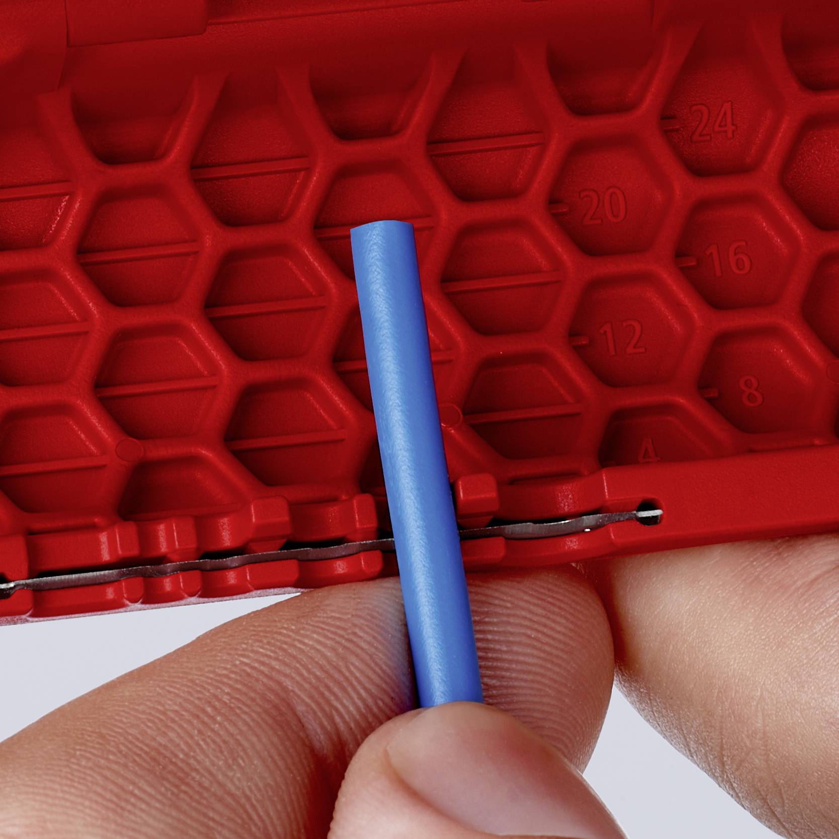 A hand holds a blue plastic rod in front of a red honeycomb structure with printed size measurements.