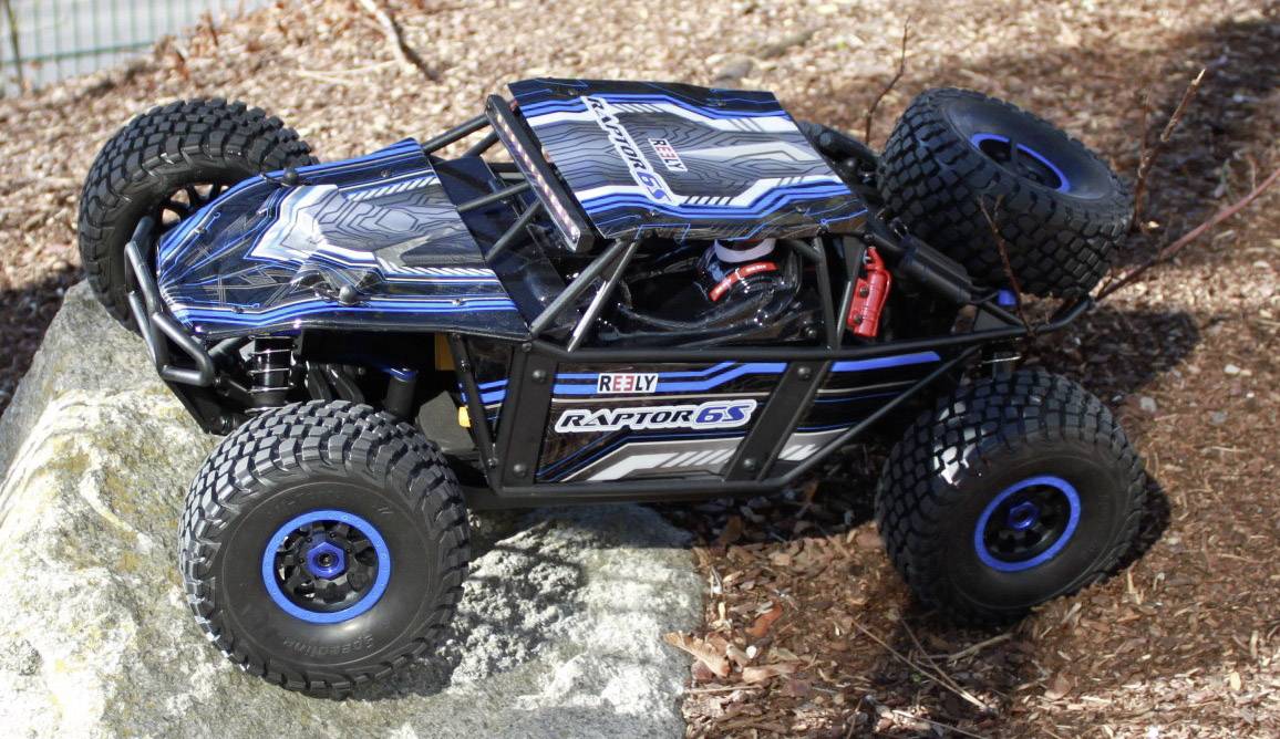 A remote-controlled car with robust tyres and a blue-black design drives over rocky terrain outdoors.