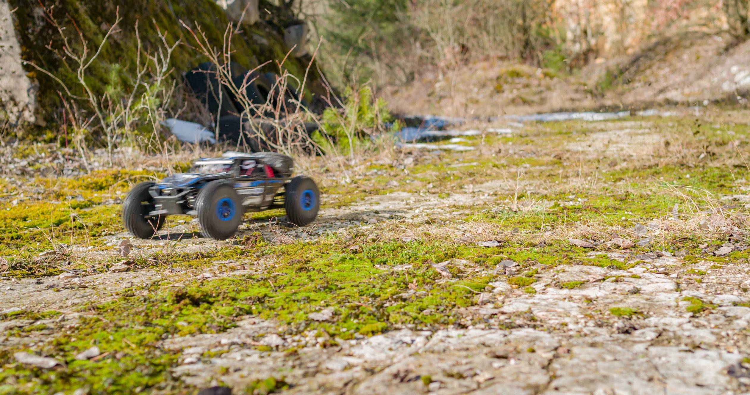 A remote-controlled car speeds across a moss-covered, rocky path in a woodland area. Trees and bushes in the background.