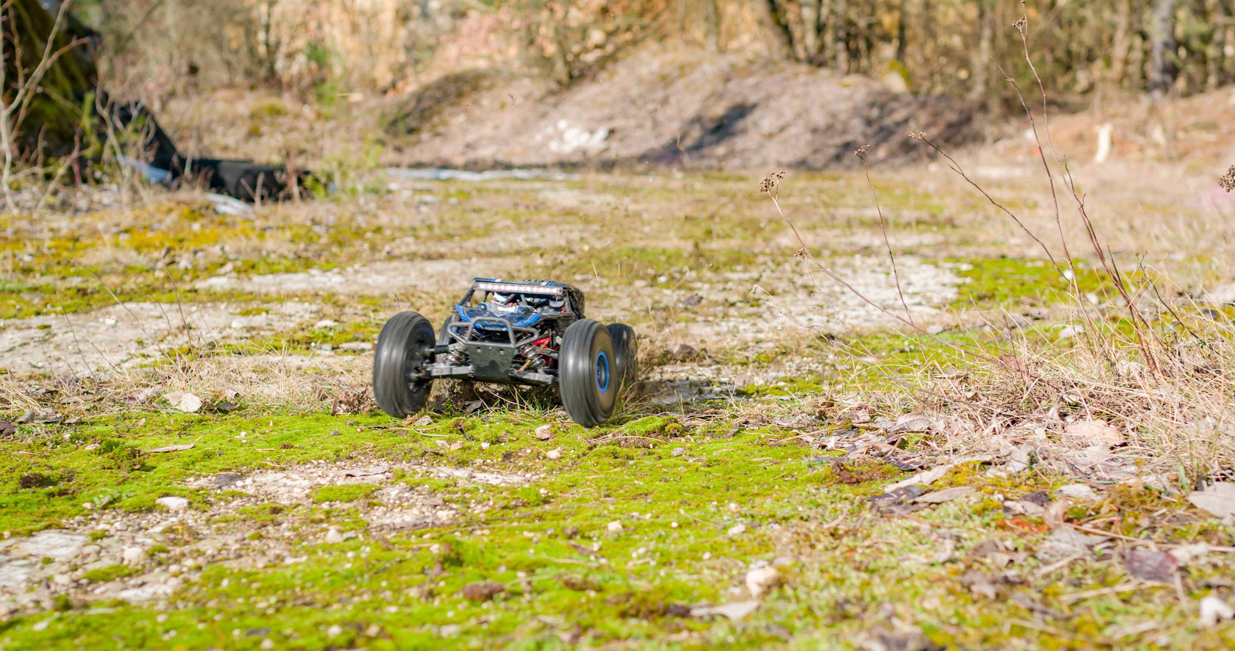 A remote-controlled car speeds along a moss-covered, rocky path in a natural setting with trees in the background.