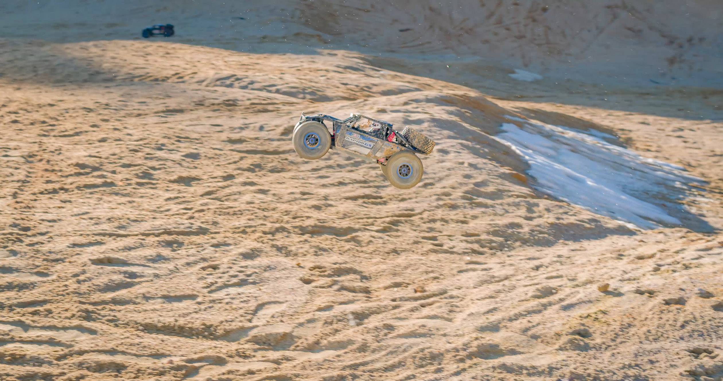 A remote-controlled car jumps through the air over a sandy dune landscape. In the background, hazy sandy ground can be seen.