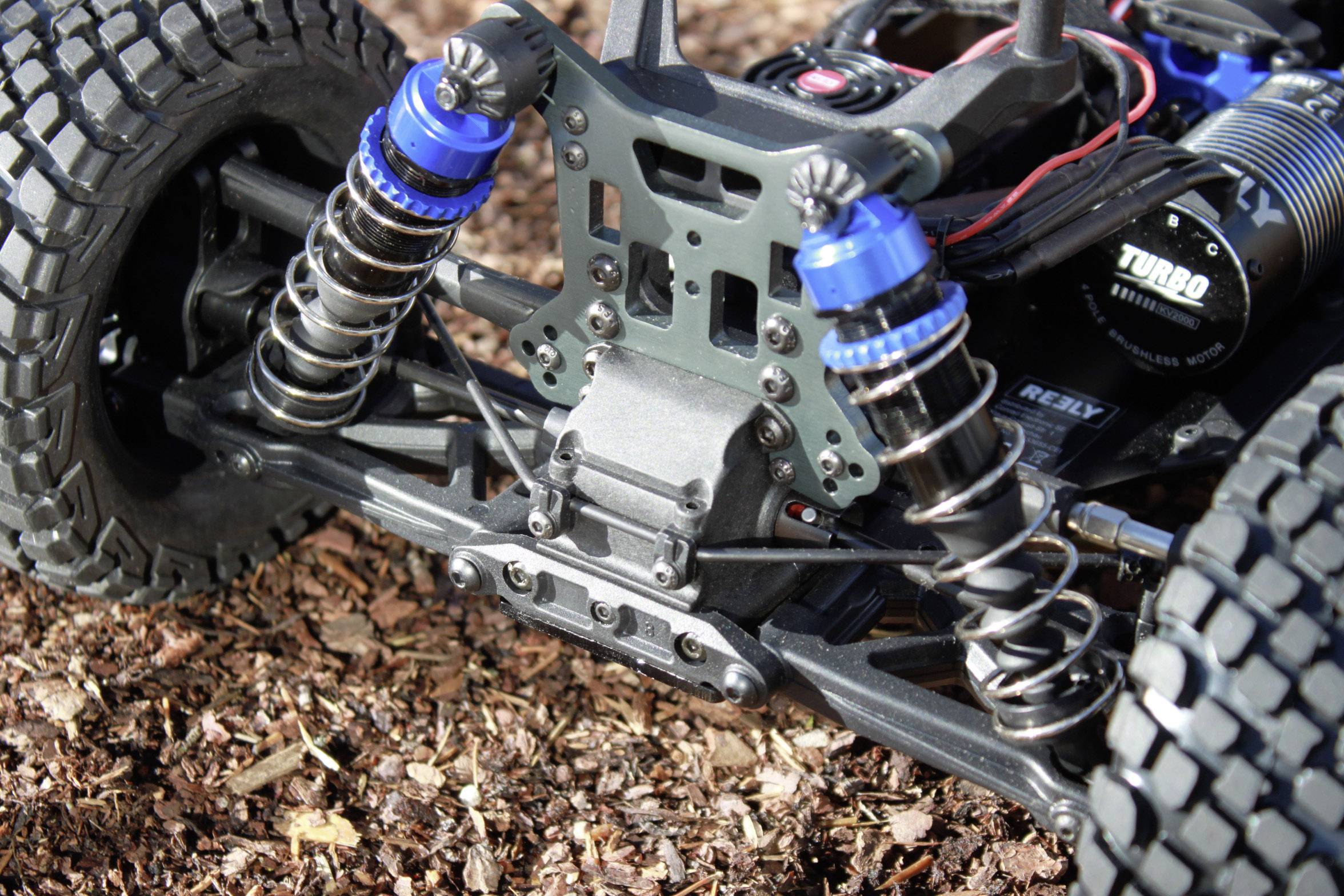 Suspension of a remote-controlled car with blue shock absorbers and chunky tyres, positioned on a surface of wood chippings.