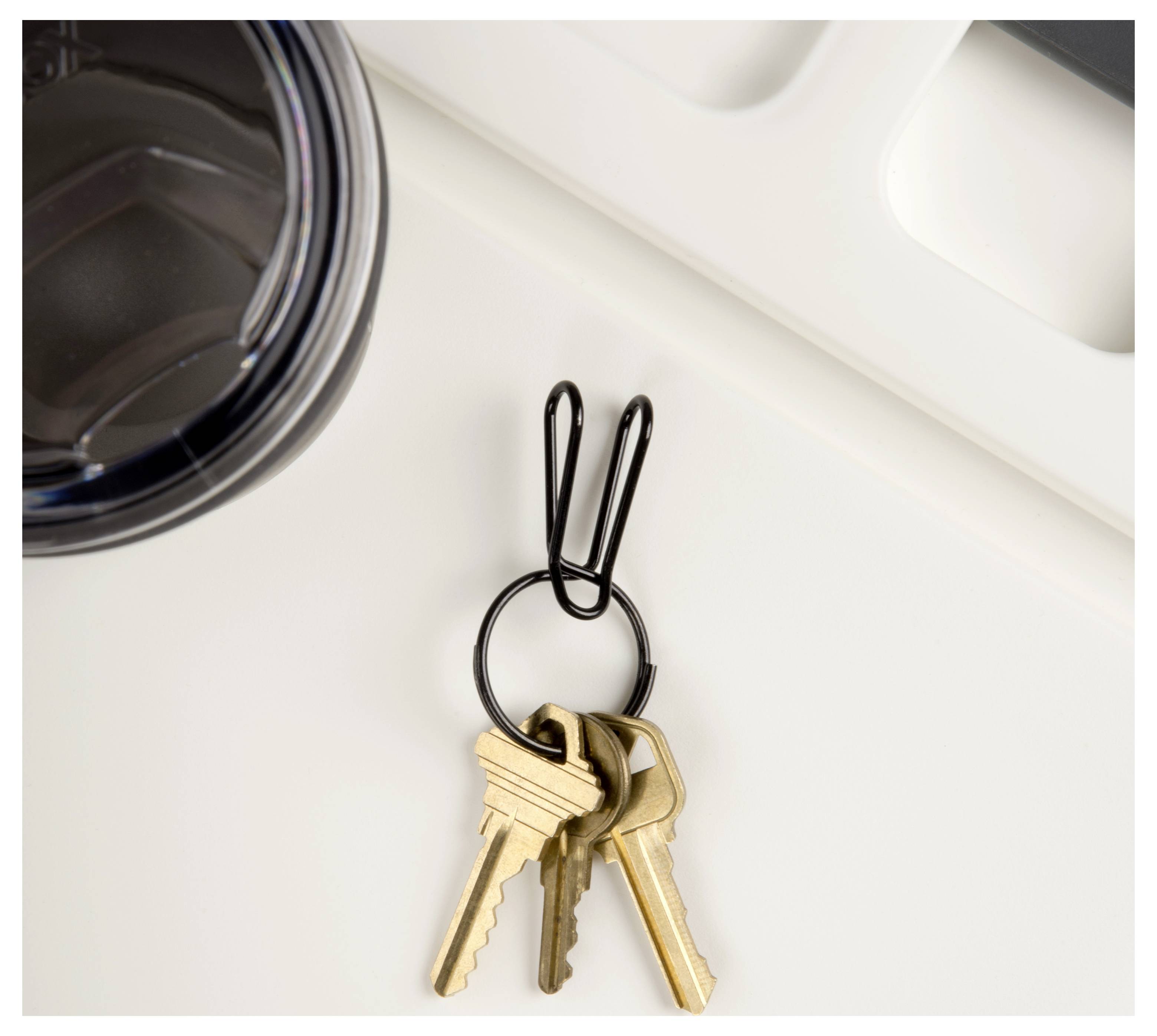 A black paperclip holds three gold keys next to a black travel mug lid on a white surface.