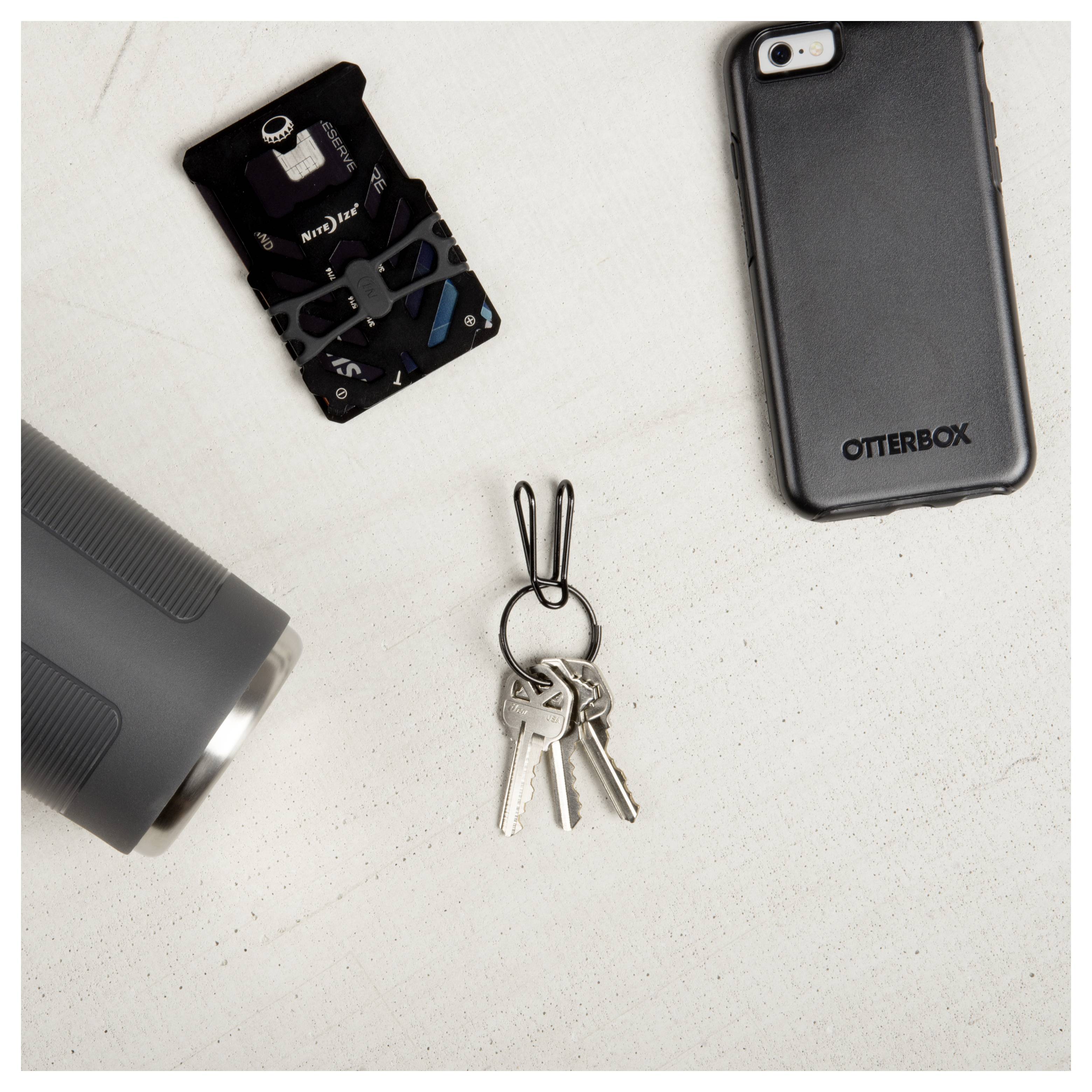Flat lay of a smartphone case, multi-tool card, metal tumbler edge, and keys with a black holder on a light surface.