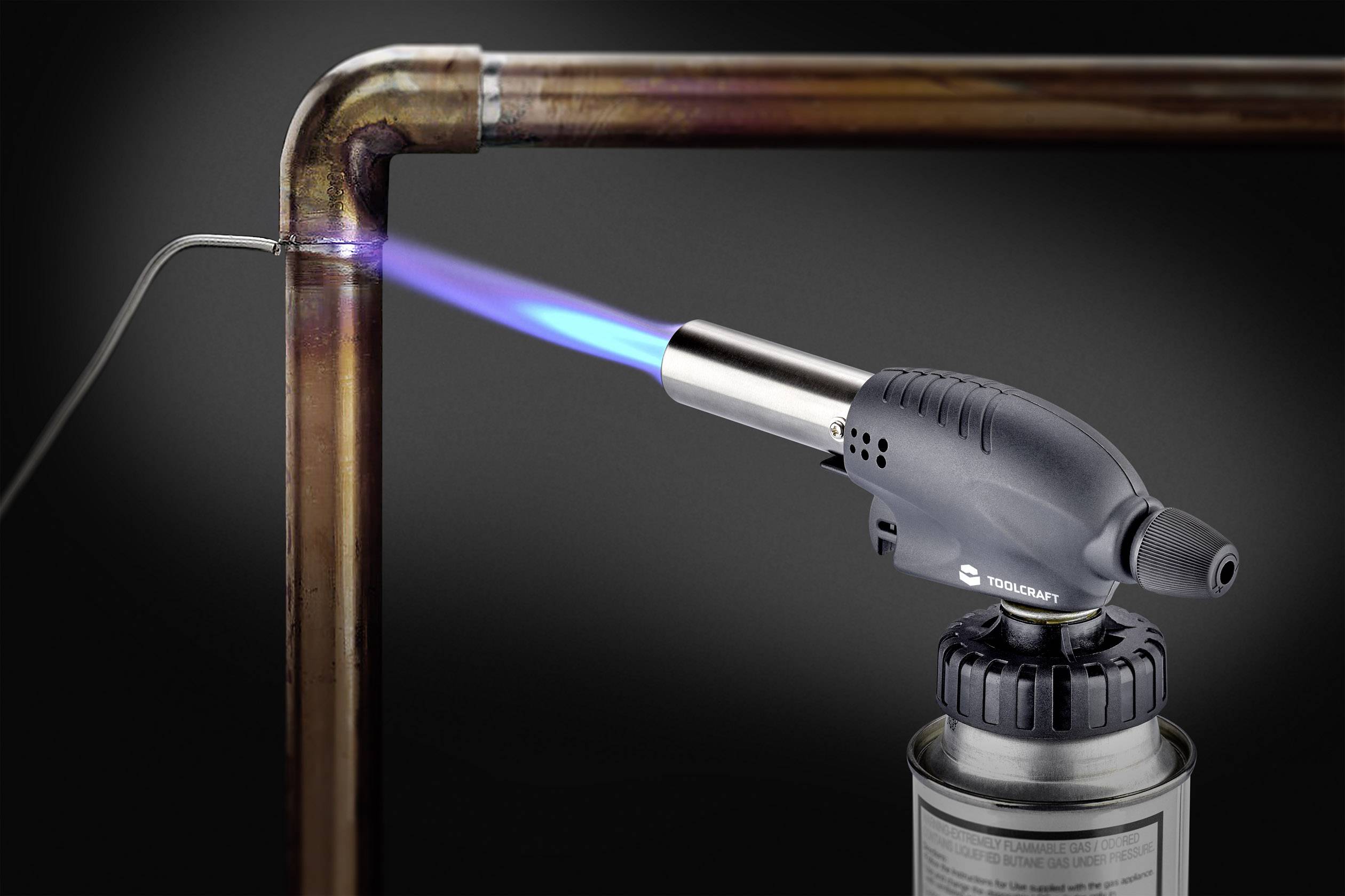 A gas burner with a blue flame is heating a copper pipe connection, presumably during a soldering task.