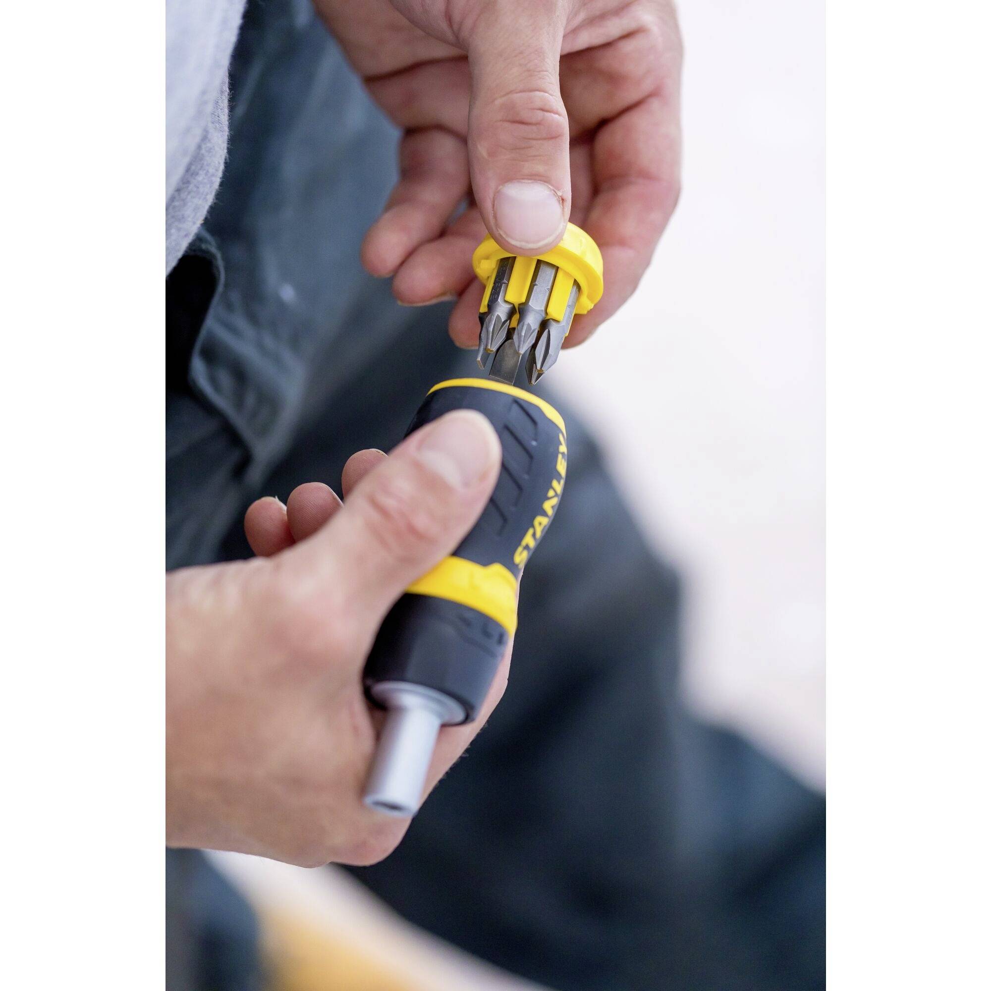 A person is holding a screwdriver with a yellow handle and an interchangeable bit set, with the focus on changing the bit.