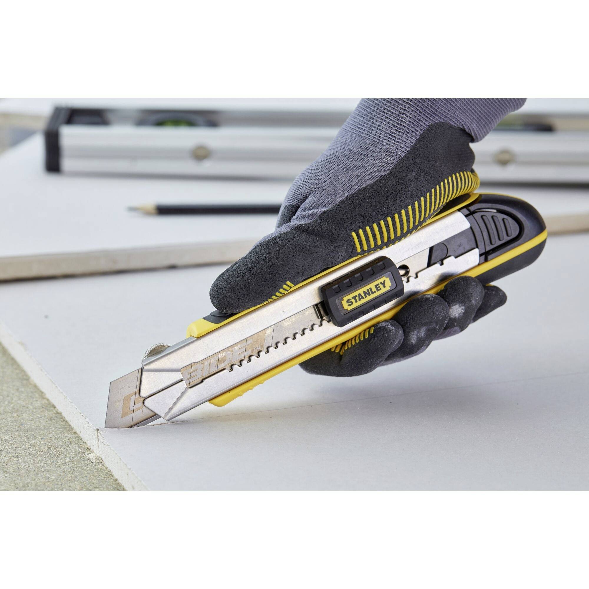 A hand wearing a glove holds a Stanley craft knife, cutting a white board. A ruler and pencil are visible in the background.