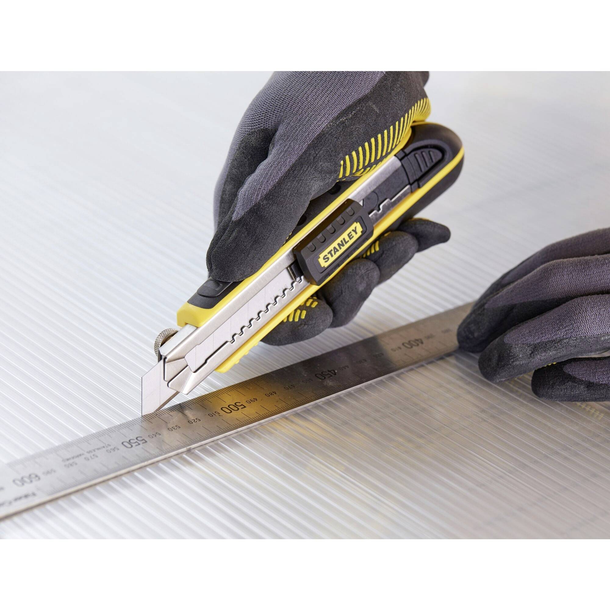 A person is cutting along a metal ruler with a yellow and black craft knife on a textured surface.