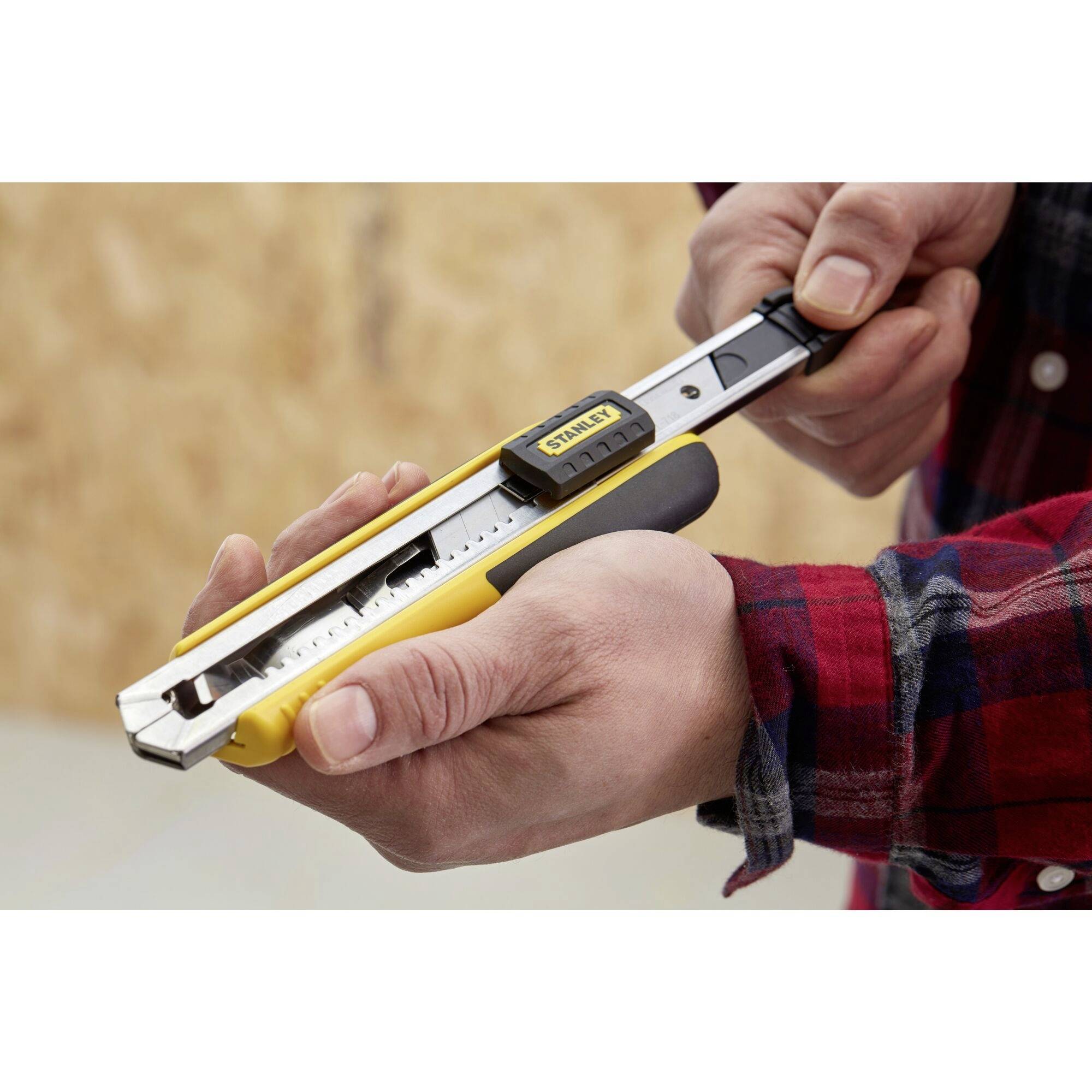 A hand in a checked shirt holds a yellow craft knife with a metal blade. Wooden panel background.