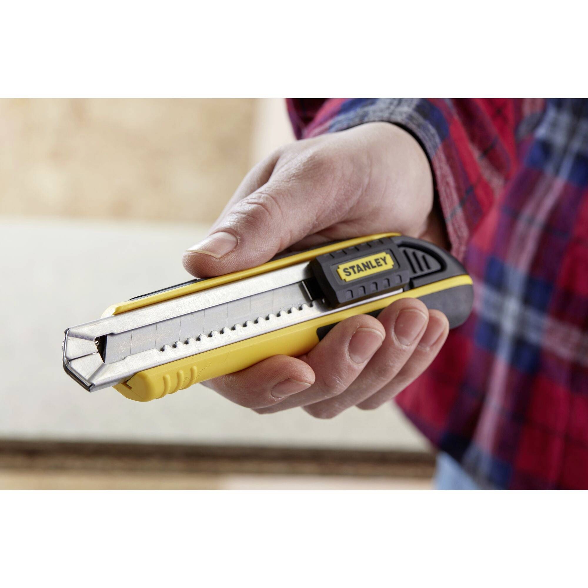 A hand is holding a yellow Stanley utility knife with an extended blade. A wooden surface can be seen in the background.