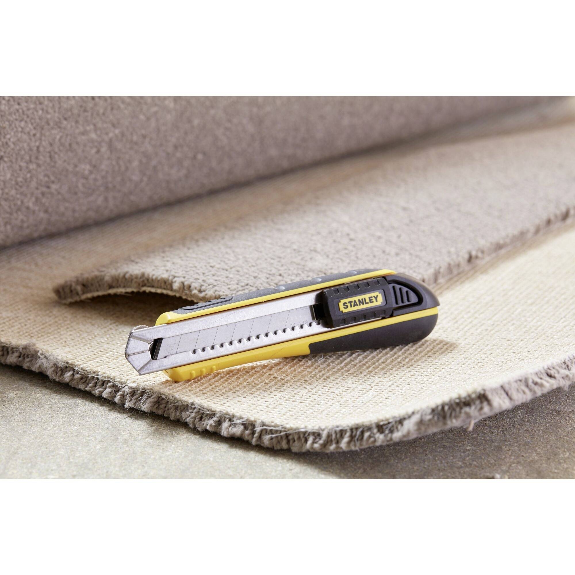 A carpet knife lies on a carpet, ready to cut. The focus is on the knife, positioned on the edge of the carpet.