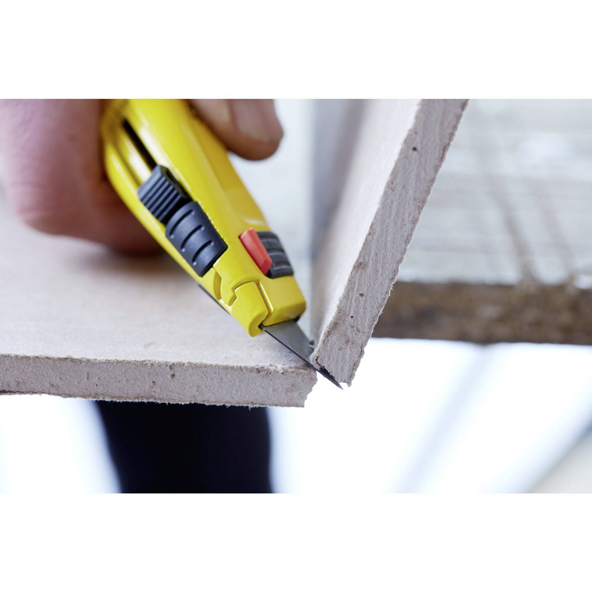 A hand is cutting a piece of plywood along the edge with a yellow craft knife.